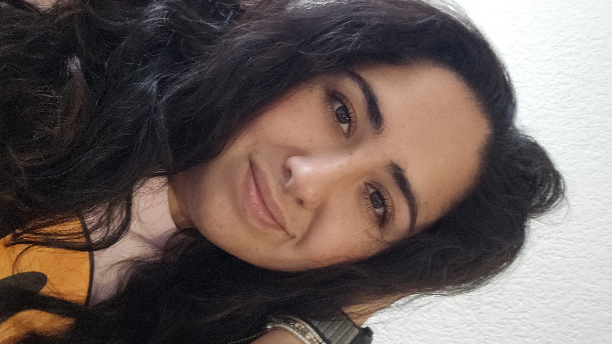 A close-up portrait of a woman with dark, wavy hair. She is smiling softly. Her eyes are brown, and she has freckles on her face. She is wearing a light pink top with an orange collar. The background is a plain white wall. The location is unknown.
