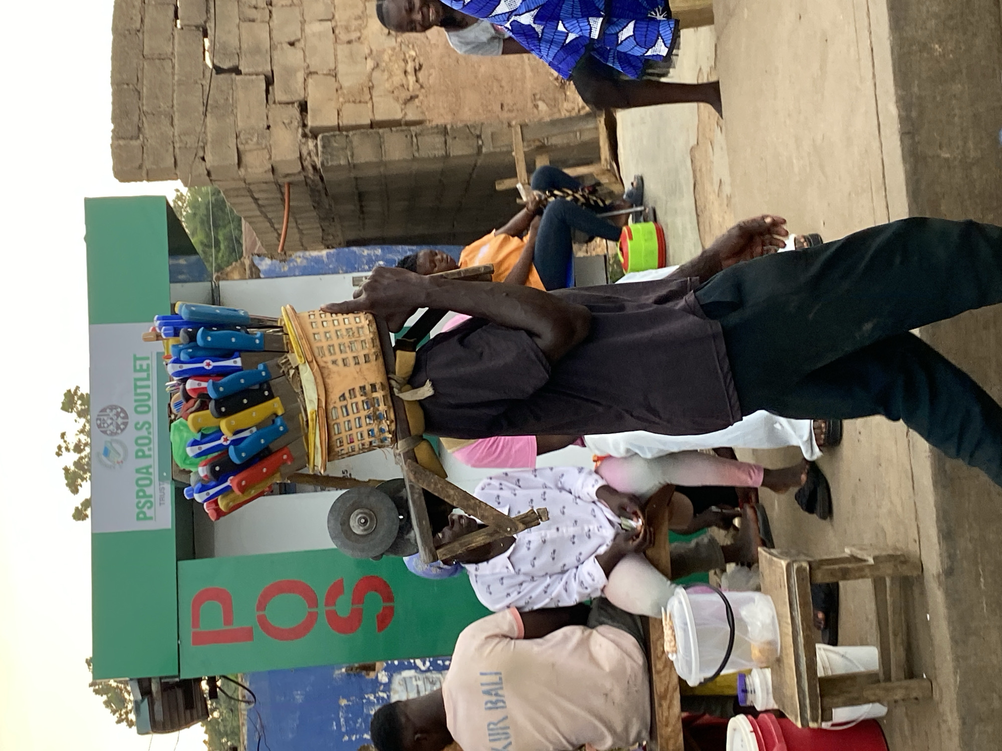 The image, taken in Bukuru, Nigeria, depicts a street scene. A knife sharpener, wearing a brown shirt and black pants, operates his mobile sharpening station, which is constructed of wood and metal and adorned with colorful knives. Customers are seated on wooden stools, waiting for their items to be serviced. A green PSPPA P.O.S Outlet sign is visible in the background. Additional pedestrians pass by, and a partially damaged brick building is situated nearby.