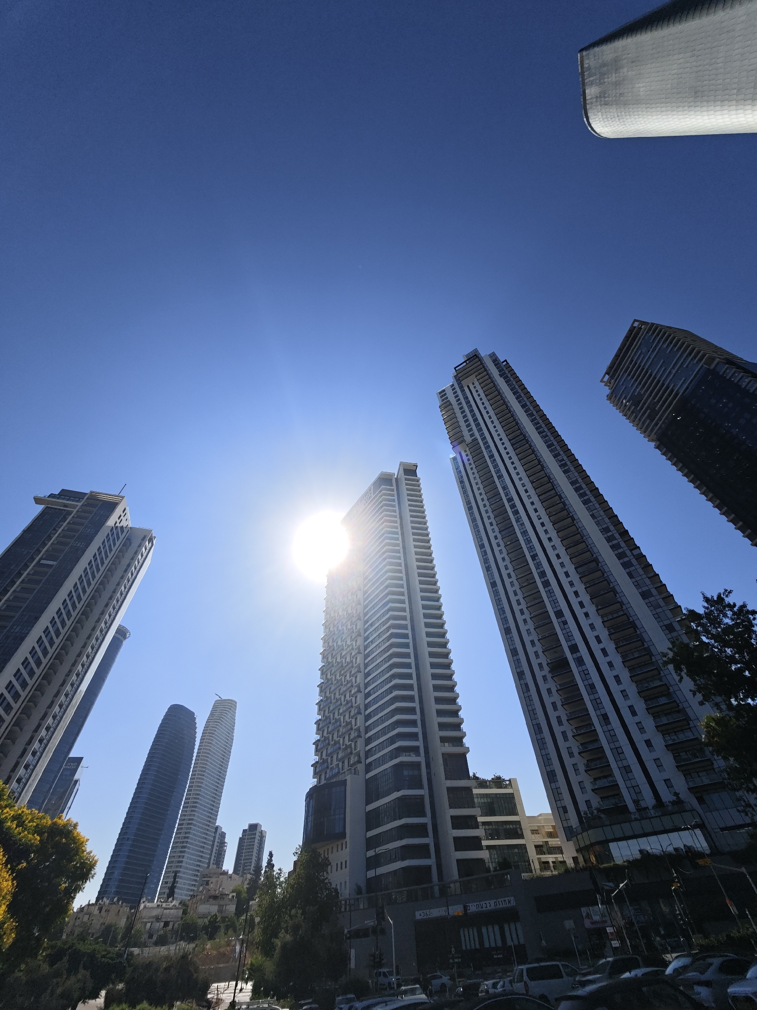 A low-angle shot captures a dense urban landscape dominated by modern high-rise buildings against a clear blue sky. The sun is bright and positioned high, creating a lens flare. Several skyscrapers of varying heights and architectural styles are visible, some with balconies and glass facades. In the foreground, trees and foliage partially obscure the lower portions of some buildings. The street level shows parked cars and some signage. The scene is consistent with a contemporary city environment, likely in Tel Aviv, Israel, given the presence of modern architecture and the style of buildings.