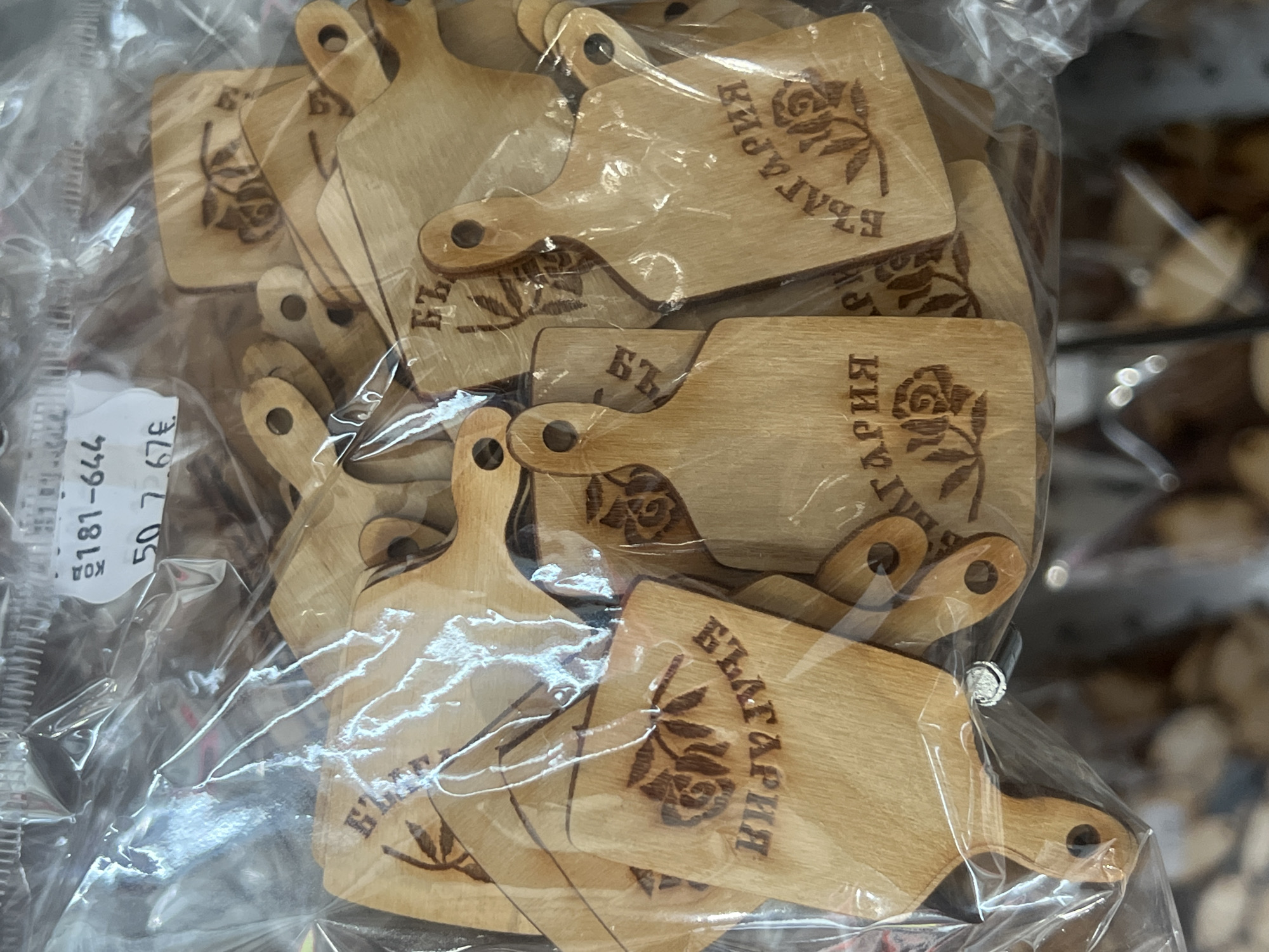 The image shows a plastic bag filled with small, light-colored wooden cutting boards. Each board has a handle with a hole. A rose and the word "България" (Bulgaria) are etched into each board. A price tag is visible on the left side of the bag, suggesting this is a retail setting. Based on the writing on the wooden boards, the location is Burgas, Bulgaria.