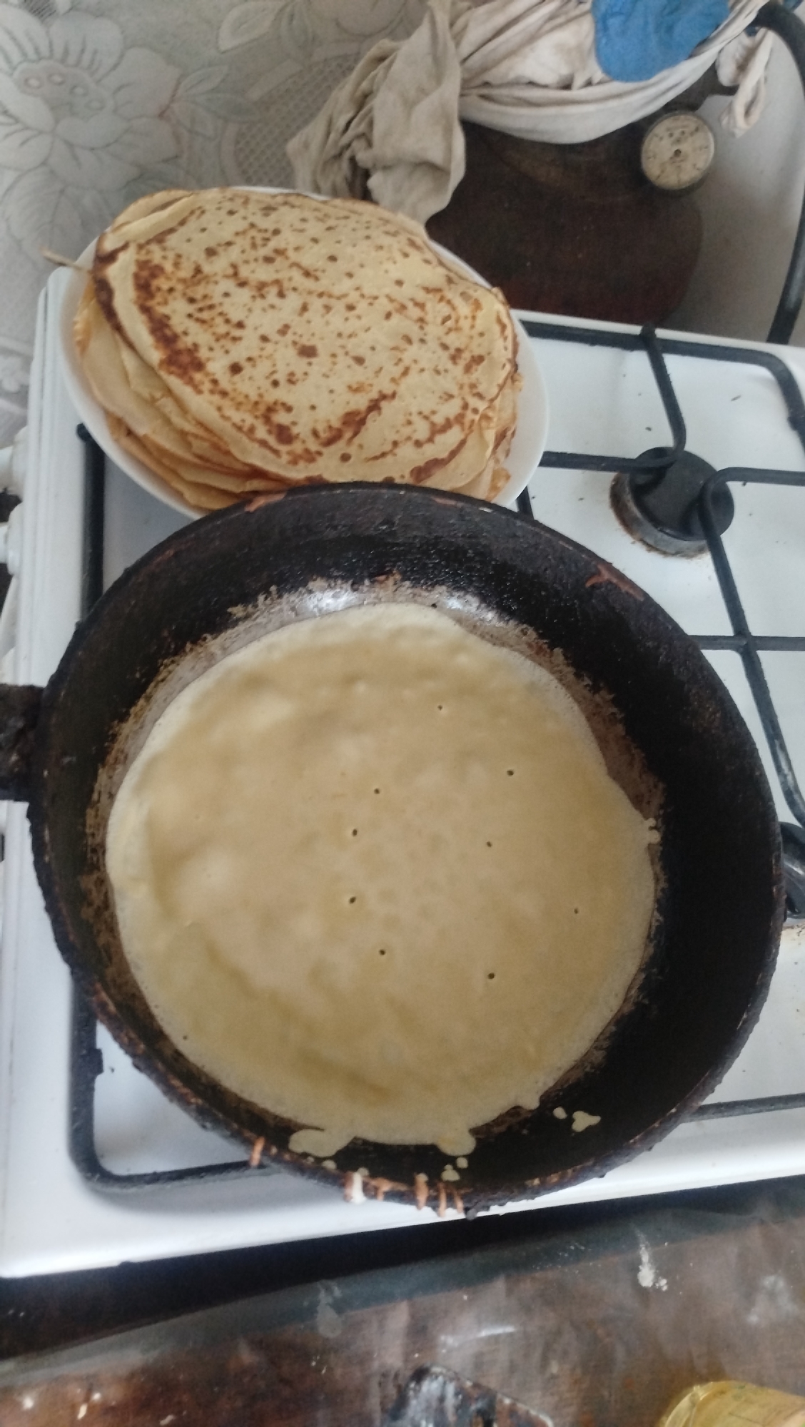 A stack of cooked crepes sits on a white plate atop a white gas stove. In the foreground, a black frying pan on the same stove contains a thin, uncooked crepe, with small bubbles forming on its surface. The stove is positioned on a countertop. In the background, a patterned curtain hangs on a wall, and to the right, a large, dark object covered with cloth is visible, with a circular gauge attached. The scene appears to be in a kitchen. The image does not provide any information to confirm its location in Kostiantynivka, Ukraine.