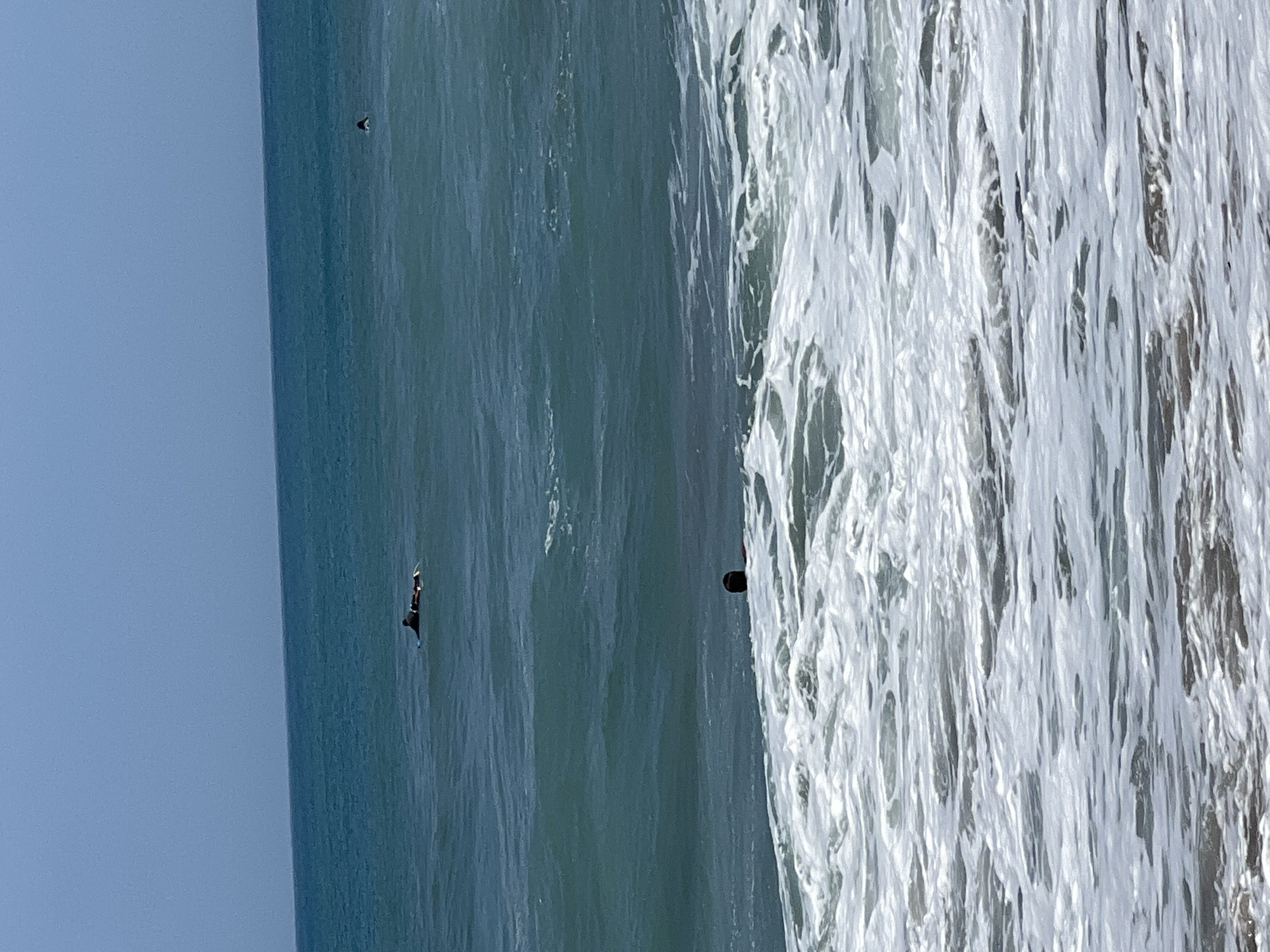 A person is lying prone on a surfboard in the ocean, with their body positioned horizontally on the board. The water is a deep turquoise, with visible ripples and waves. In the foreground, white foam from a breaking wave washes towards the viewer. The sky is clear and a pale blue. Two other surfers are visible in the distance. One is also on a surfboard, while the other is further out and only their head is visible above the water. The scene appears to be at Kuta Beach, Bali, Indonesia.