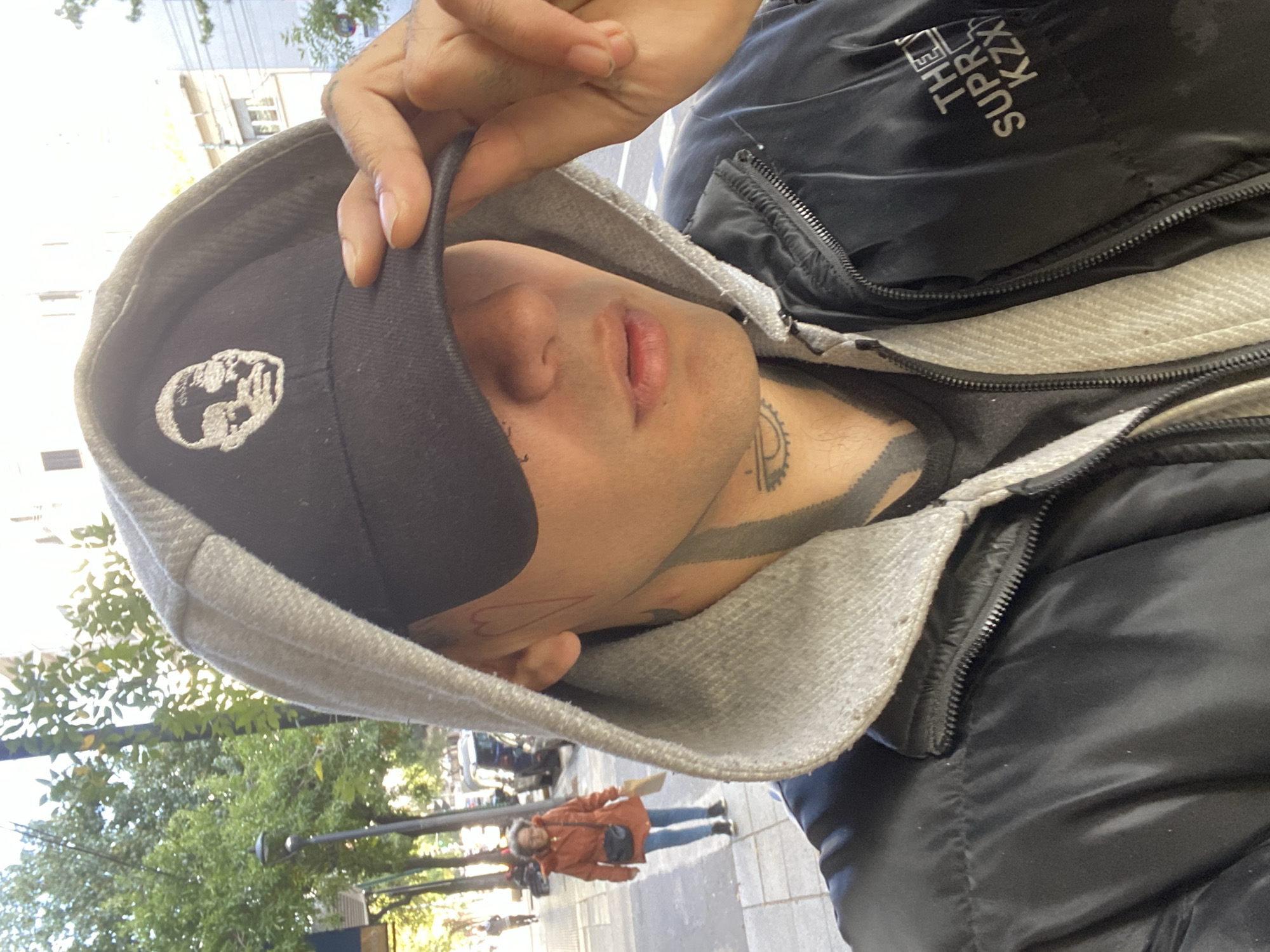 A selfie shows a man wearing a black baseball cap with a white skull-like design, a grey hooded sweatshirt, and a black jacket. The man has tattoos on his neck and face. In the blurred background, people are walking on a sidewalk in a cityscape setting. The location is likely in Buenos Aires, Argentina.