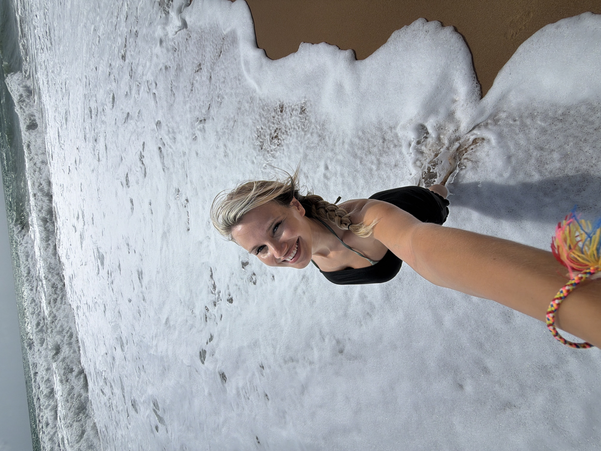 A woman with blonde hair in a braid is taking a selfie at the beach. She is wearing a black tank top and is smiling at the camera. The ocean waves are crashing on the shore, creating a lot of foam. The sand is wet and brown. The sky is overcast. The location is Mai Khao, Thailand.