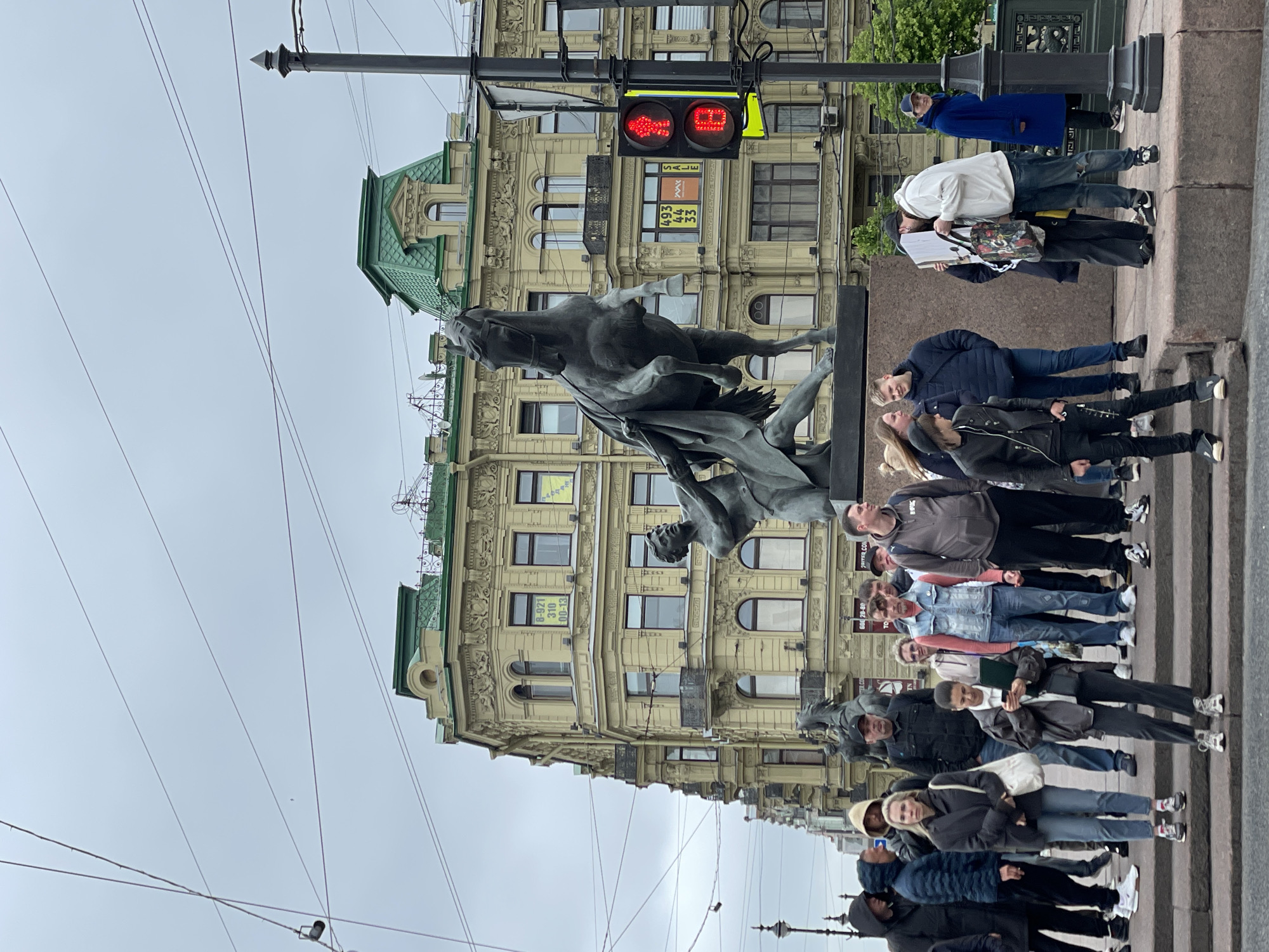 The image shows a crowd of people standing near a statue of a horse and rider in Saint Petersburg, Russia. The statue is bronze and depicts a rider struggling with a horse. The crowd is a mix of ages and genders, and they are all looking at the statue. In the background, there is a building with ornate architectural details. A traffic light is visible, showing a red pedestrian signal. The weather appears overcast, with a gray sky. Overhead wires are visible.
