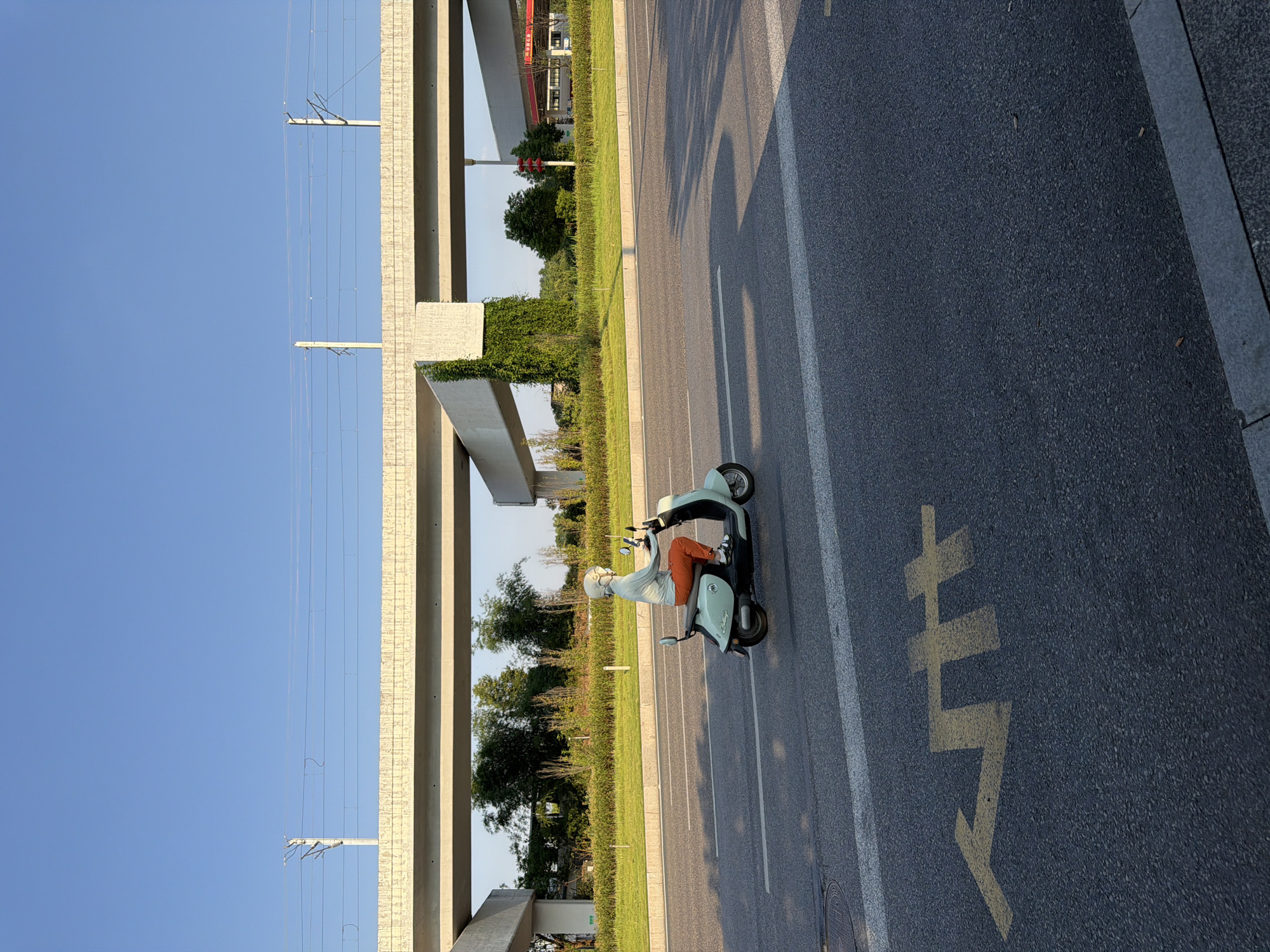 A person rides a mint green scooter on a paved road in Jiangjin, China, under a clear blue sky. The scooter rider is wearing a light blue hooded jacket, a white helmet, and orange pants. The scooter has a black seat and a silver backrest. The road has a yellow arrow painted on it, indicating a turn. In the background, a concrete overpass stretches across the road, with power lines running above it. Green vegetation, including trees and bushes, grows alongside the road and under the overpass. A gas station with a red roof is visible in the distance. The lighting suggests it is daytime, possibly late afternoon, given the long shadows cast on the road.