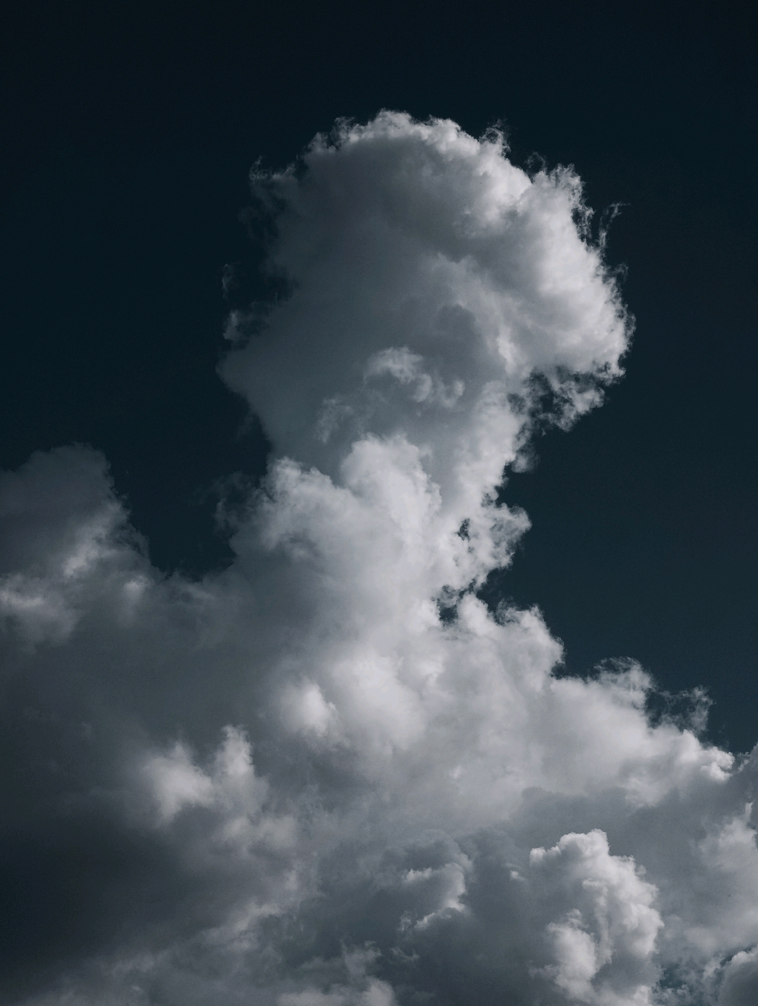 An isolated shot captures a dramatic, dark blue sky filled with voluminous, billowing clouds. The clouds are predominantly white and gray, with a stark contrast between the illuminated tops and shadowed undersides, suggesting a dynamic interplay of light and shadow. The overall impression is one of moody, atmospheric grandeur.