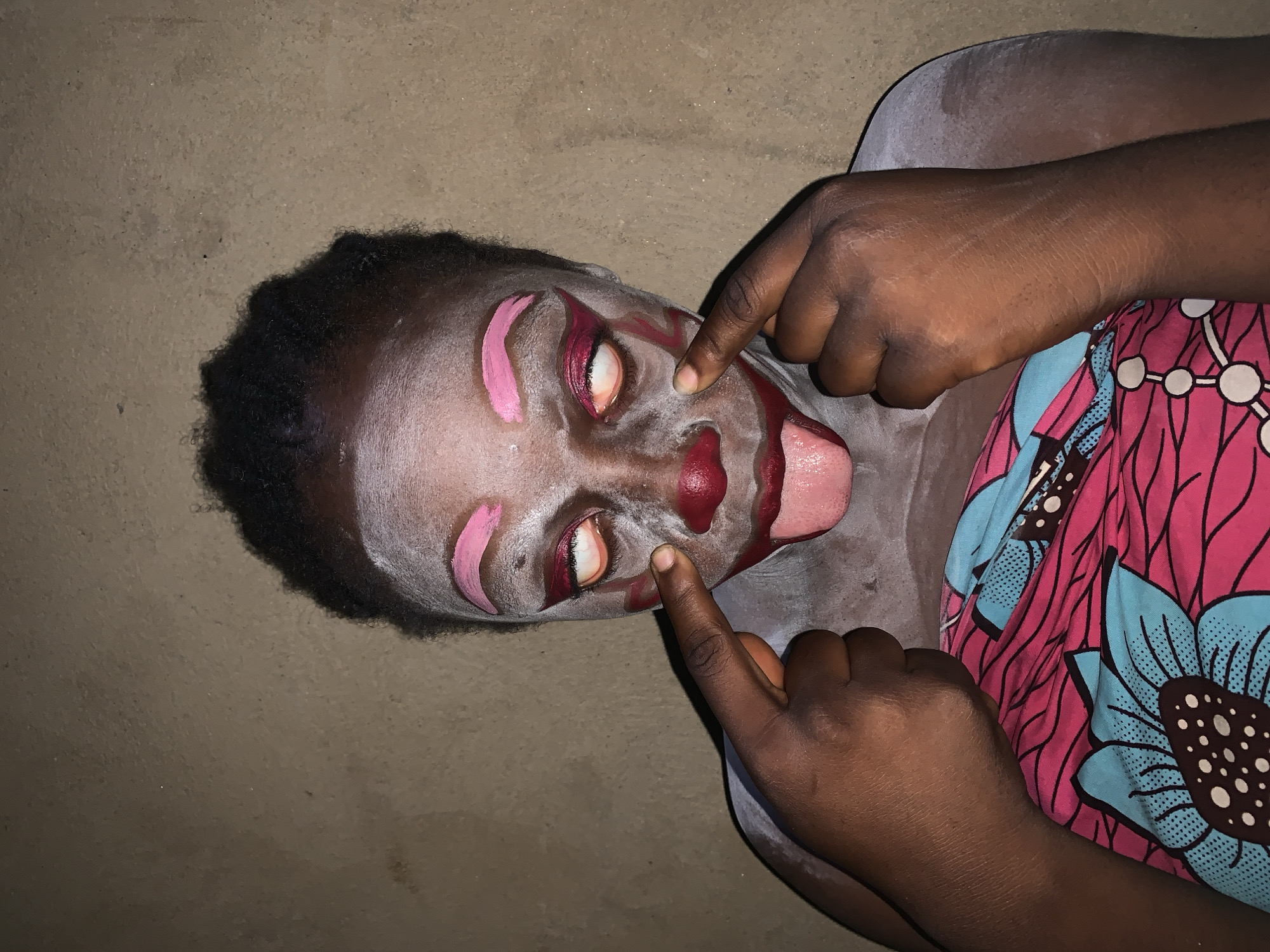 The image shows a close-up of a person's face with clown-like makeup. The person has white face paint, pink eyebrows, red accents around the eyes, a red nose, and red lips with their tongue sticking out. Their eyes are open, showing the whites of their eyes, and they are pointing at their cheeks with their fingers. The background is a textured, earth-toned wall, consistent with the environment found in Agban, Nigeria. The person is wearing a brightly colored fabric with a pink background and blue and black floral patterns.