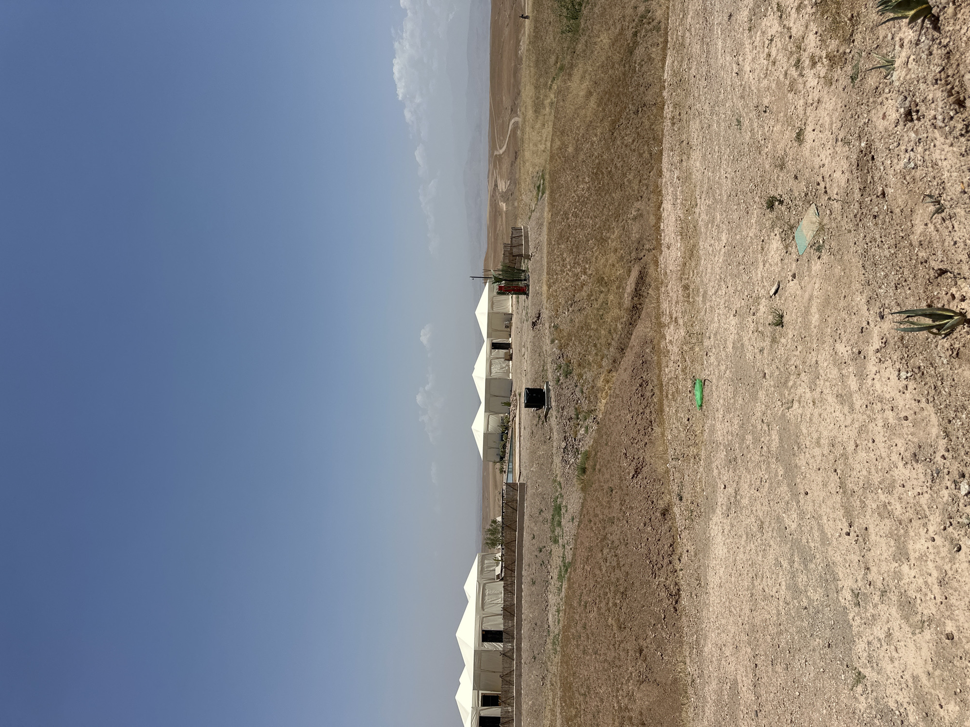 The image depicts a series of white tents in a desert-like environment, possibly a luxury campsite, under a clear blue sky. The terrain is arid and consists of dirt and sparse vegetation. In the background, there are hills or mountains under a hazy sky. The location is near Sidi Badhaj, Morocco. There are no people visible. Some tents have visible openings, suggesting they are inhabitable. The image is rotated 90 degrees counterclockwise. A few agave plants are visible in the foreground. There are green and blue objects on the ground.