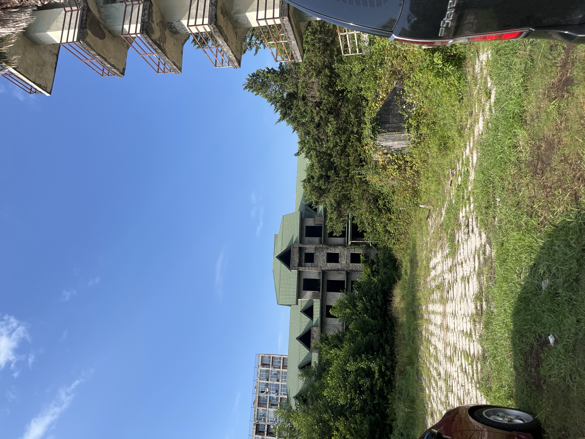 This is a medium shot photograph of an abandoned building complex in Ureki, Georgia. In the foreground, a portion of a dark-colored minivan is visible on the right, and a red car is parked on the left. A paved area with interlocking concrete blocks is visible between the cars and the buildings, covered with patches of green grass.
The central focus is a multi-story building with a green tiled, pitched roof. The building appears to be under construction or abandoned, with unfinished concrete walls and dark, empty window openings. The roof features multiple gables and dormer windows. To the left of this building, a taller, more modern apartment building with numerous windows and balconies is visible in the background.
To the right of the main building, another concrete structure with multiple balconies is partially visible. These balconies have rusty metal railings and show signs of wear and graffiti. Lush green trees and vegetation surround the buildings, with some vines growing on a weathered wooden structure to the right of the central building. The sky is clear blue with a few wispy white clouds. The overall impression is one of urban decay amidst natural overgrowth.