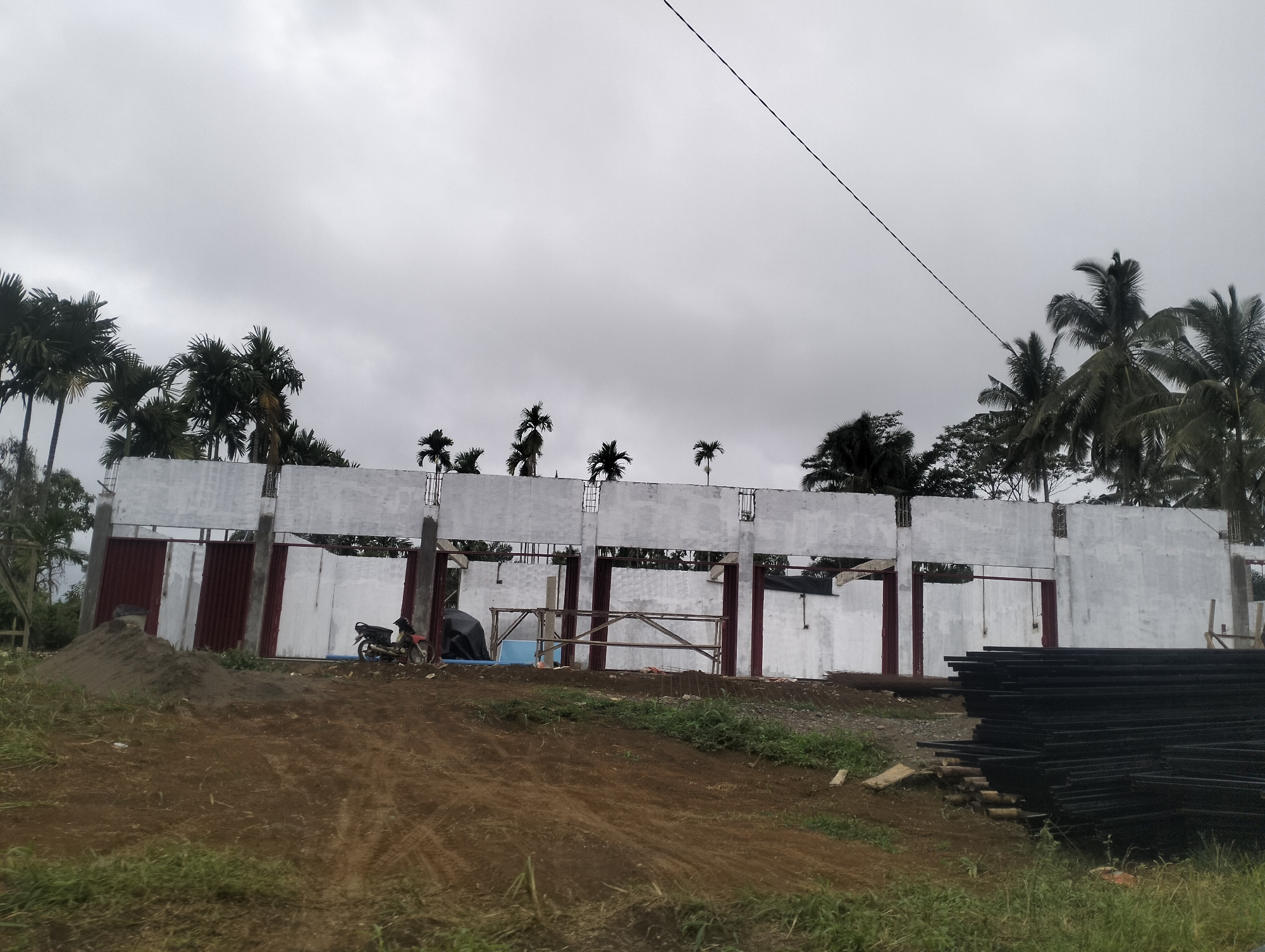 The image depicts an outdoor construction site under a cloudy sky. In the background, a partially constructed building with white walls and visible structural elements is present. Red corrugated metal panels are installed in some of the openings. Several palm trees are visible behind and above the structure. In the foreground, a pile of dark metal bars is neatly stacked on the right side of the image. A motorcycle is parked near the building, and a mound of dirt is situated to the left. The ground is covered with rough terrain, dirt, and some patches of green grass. The overall scene suggests ongoing construction in a tropical or subtropical location.