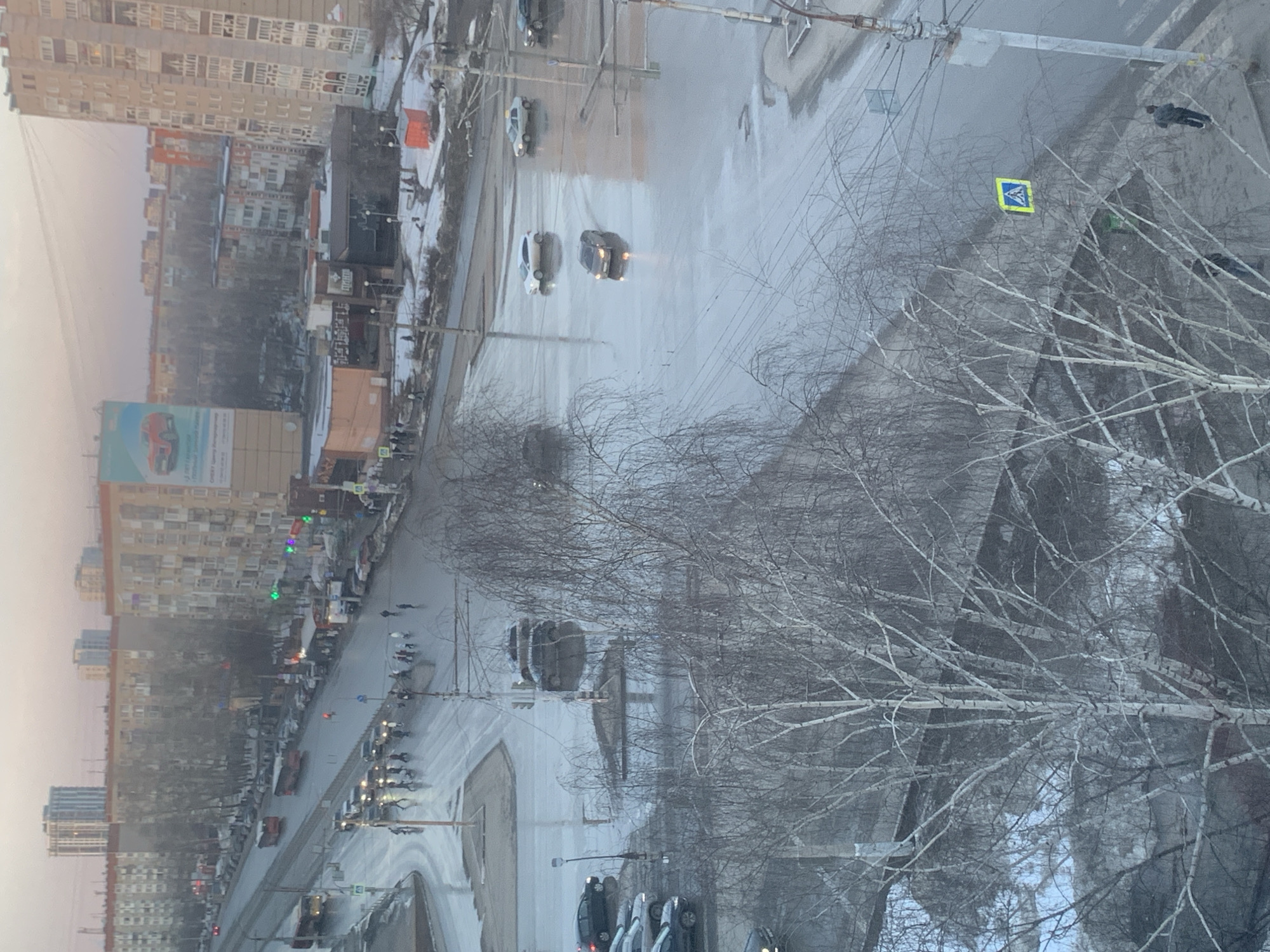 The image depicts an elevated view of a busy urban intersection in Izhevsk, Russia, under an overcast sky. The roads are extensively wet and reflective, with visible puddles, suggesting recent precipitation or significant snowmelt. Patches of dirty snow and ice are present on unpaved areas and alongside sidewalks.
**Roads and Traffic:**
A wide, multi-lane main road forms an intersection in the mid-ground. Vehicular traffic includes a dark sedan with illuminated headlights moving through a wet section, a white SUV, and a light-colored vehicle resembling a taxi further right. On the left side of the main road, two red articulated buses are in motion. Multiple parked cars line the street and adjacent parking areas. Traffic lights displaying both red and green signals are active at the intersection.
**Pedestrians:**
A group of approximately 15 to 20 individuals, predominantly dressed in dark outerwear, are actively crossing the main intersection at a marked crosswalk. Additional pedestrians are visible on sidewalks and near buildings. A blue and yellow pedestrian crossing sign is affixed to a pole in the lower right foreground.
**Buildings and Urban Environment:**
The background features numerous multi-story residential apartment buildings, consistent with Soviet-era architecture. At street level, commercial buildings are visible, including one with a large vertical billboard advertising a red Chery Tiggo vehicle. Another building has a dark facade and signage in Cyrillic. A bright orange, cube-shaped structure stands on a snowy patch of ground to the right of the intersection.
**Vegetation and Infrastructure:**
Numerous bare deciduous trees, identifiable as birch trees by their light-colored bark, dominate the foreground and line the streets, indicating a dormant season. Overhead power lines, utility poles, and streetlights are visible throughout the scene.