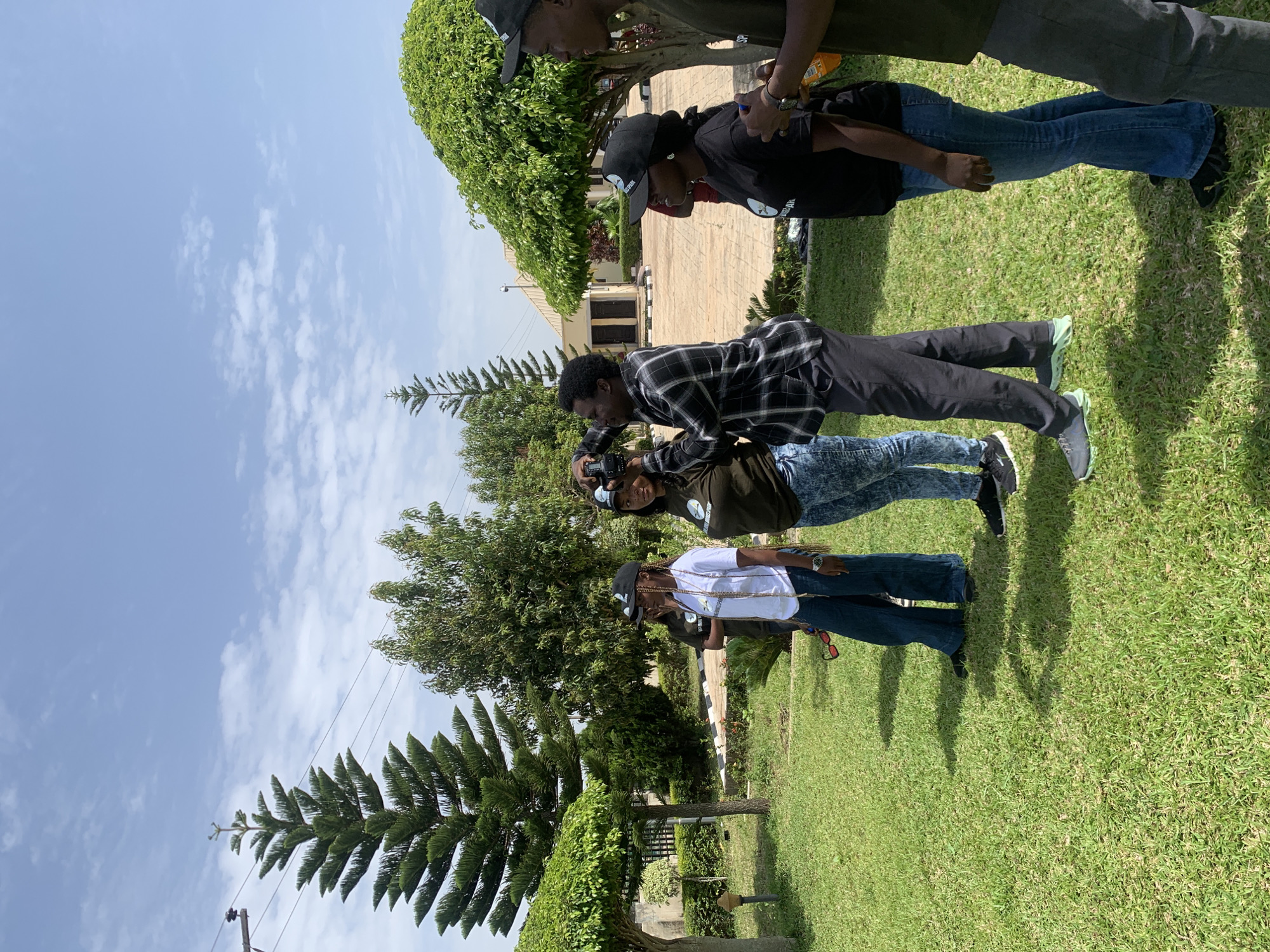 This media file depicts a group of young people outdoors on a sunny day, likely in Jos, Nigeria. The scene is a grassy area with trees and some buildings in the background. The time of day appears to be late morning or early afternoon, suggested by the bright sunlight casting distinct shadows. The weather seems pleasant, with a clear blue sky dotted with white clouds.
There are at least four individuals visible, all dressed casually in t-shirts, jeans, and baseball caps. One person, wearing a checkered shirt and dark pants, is holding a camera and appears to be taking a photograph or video of another person. This suggests an informal gathering, possibly a photoshoot or a casual hangout where someone is documenting the event.
One of the people is wearing a black t-shirt with white text that seems to be a logo or emblem. Another person, wearing a white t-shirt, also has some visible designs or text on their clothing, though it is difficult to read clearly. One of the baseball caps has the word "PINK" visible on it.
The overall mood appears relaxed and casual. The individuals are engaged with each other and the activity at hand, suggesting a friendly and comfortable atmosphere. The presence of lush green grass and mature trees indicates a well-maintained outdoor space, possibly a park, a residential compound, or an event venue.