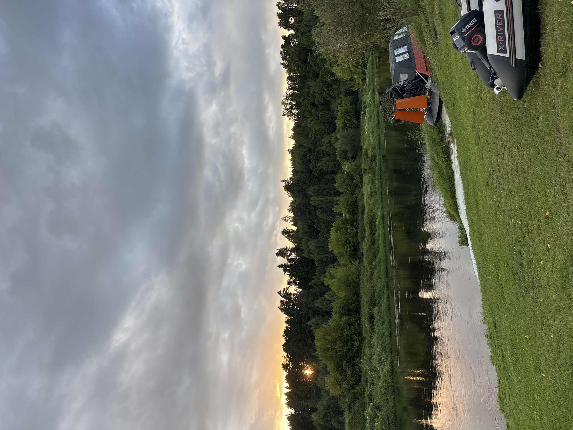 This image depicts a serene riverside scene in Zvenigorod, Russia, likely during sunset. The foreground features a grassy bank with two inflatable boats parked on it. One boat is larger, red and black with an orange shroud over a fan, suggesting it's an airboat or hovercraft. The other is a smaller, black and white inflatable boat with an outboard motor labeled "YAMAHA" and "X-RIVER".
The middle ground is dominated by a calm river reflecting the surrounding landscape and the soft, diffused light of the setting sun. The riverbank on the opposite side is densely covered with a variety of trees and vegetation, forming a lush green forest.
The background is a sky filled with dramatic, grey clouds, through which the warm, golden light of the setting sun is breaking. The sun is partially visible, casting a gentle glow through the trees on the horizon. The overall atmosphere is peaceful and natural, highlighting the beauty of the Russian countryside.