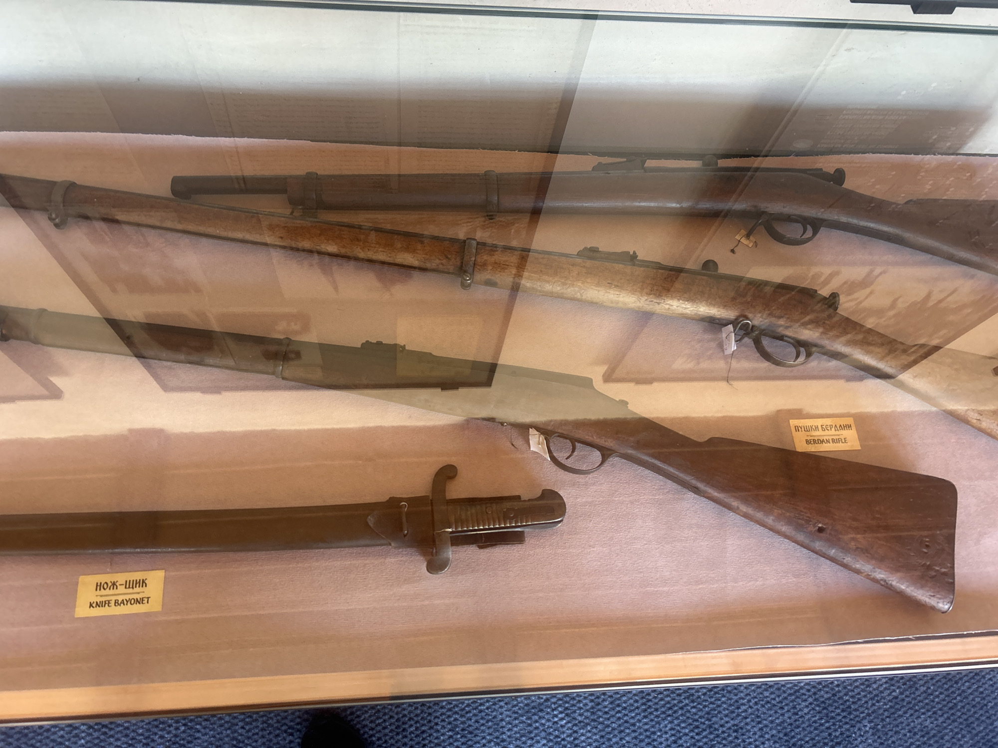 This image depicts a display case containing several antique firearms, likely in a museum or historical exhibit. The firearms are arranged vertically and appear to be rifles with bayonets attached to at least one of them. One rifle is partially visible on the left, showing its wooden stock and the bayonet mounted on the barrel. To the right, two more rifles are visible, their wooden stocks and metal components clearly discernible.
A yellow label is attached to the wooden stock of the rifle on the left, with text that appears to be in Cyrillic script and the English words "KNIFE BAYONET". Another yellow label is present on the wooden stock of the rifle on the far right, with Cyrillic script and the English words "BERDAN RIFLE". The background is a textured, light-colored surface. The glass of the display case creates reflections, making some details appear layered or slightly obscured. The context provided indicates this display is located in the city of Belogradchik, Bulgaria.