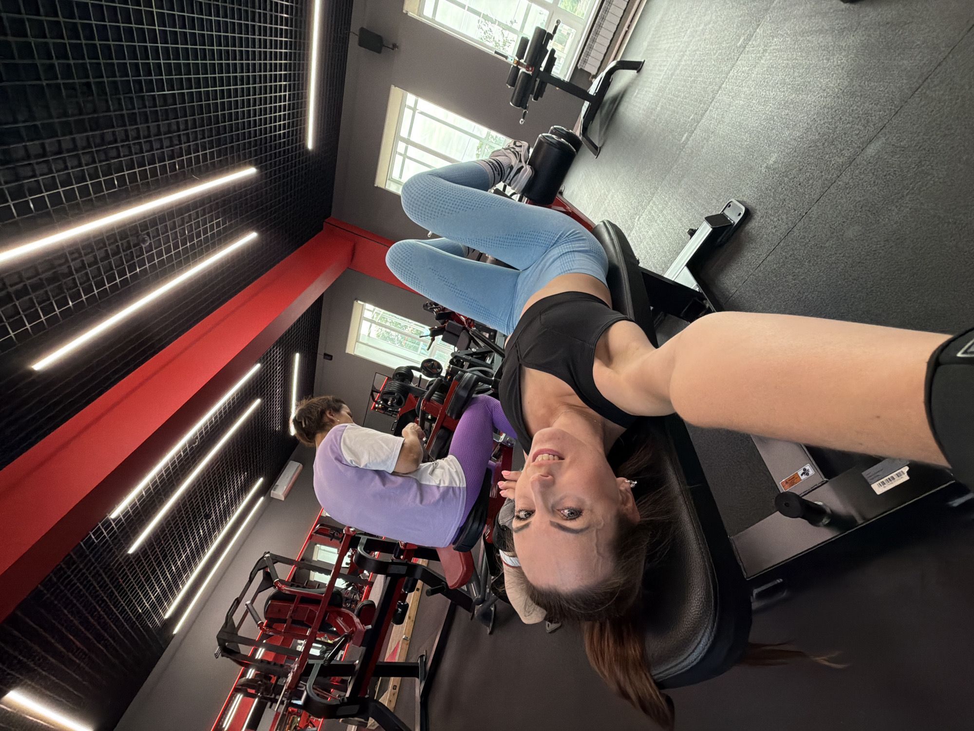 The image depicts the interior of a modern gym, possibly located in Birobidzhan, Russia. In the foreground, a woman is lying on her back on a weight bench, holding a camera for a selfie. She is wearing a black sports bra and blue leggings. Her head is tilted back, and she is smiling at the camera. In the background, another person is seated on a weight machine, also wearing athletic attire. The gym features a black grid ceiling with modern LED lighting and red accents. Various weight machines and gym equipment are visible throughout the space. Large windows allow natural light to enter the gym. The overall environment suggests a functional fitness facility.