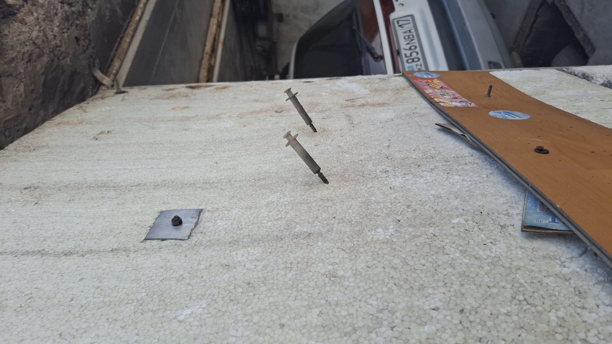 The image shows a close-up, angled view of a textured, light-colored wall, possibly made of styrofoam insulation, in an outdoor or semi-outdoor setting. Two syringes are stuck into the wall, with their needles pointing inwards and their plungers facing outwards. To the left of the syringes, a small, square metal plate with a bolt is also affixed to the wall. In the lower part of the image, a wooden plank with stickers and what appears to be a book or magazine peeking out from underneath is leaning against the wall. In the background, out of focus, a portion of a car with a license plate that reads "KZ 856NBA 17" is visible. The overall environment appears to be utilitarian and somewhat dilapidated. Without further context or specific landmarks visible in the image, it is not possible to determine its exact location within the city of Shymkent, Kazakhstan.