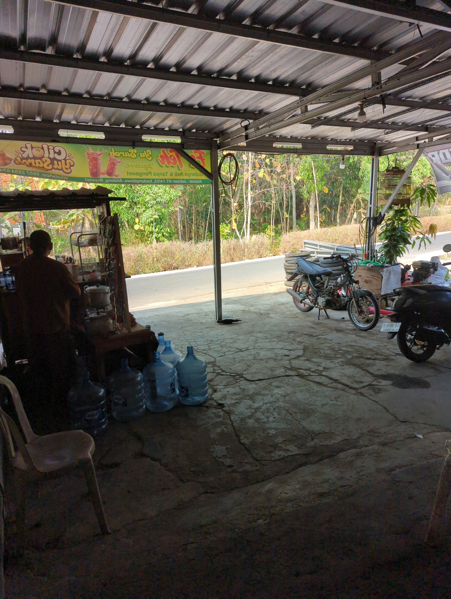 The image depicts an outdoor covered area, likely a workshop or roadside stall, in Kedungdowo, Indonesia. A person, seen from behind and silhouetted, is standing at a counter, possibly preparing drinks. Various items are arranged on shelves behind them, including bottles and containers. In the foreground, there are several large water cooler jugs.
To the right, two motorcycles are parked. One is a dark-colored scooter, and the other is a more traditional motorcycle with exposed engine parts, suggesting it might be undergoing repairs or maintenance.
The ground is paved with concrete, showing some cracks and discoloration. Beyond the covered area, there is a road and a backdrop of lush green trees and vegetation, indicating a semi-rural or natural environment. A bird cage is hanging from a post on the right side of the image. The overall impression is of a functional, open-air establishment.
