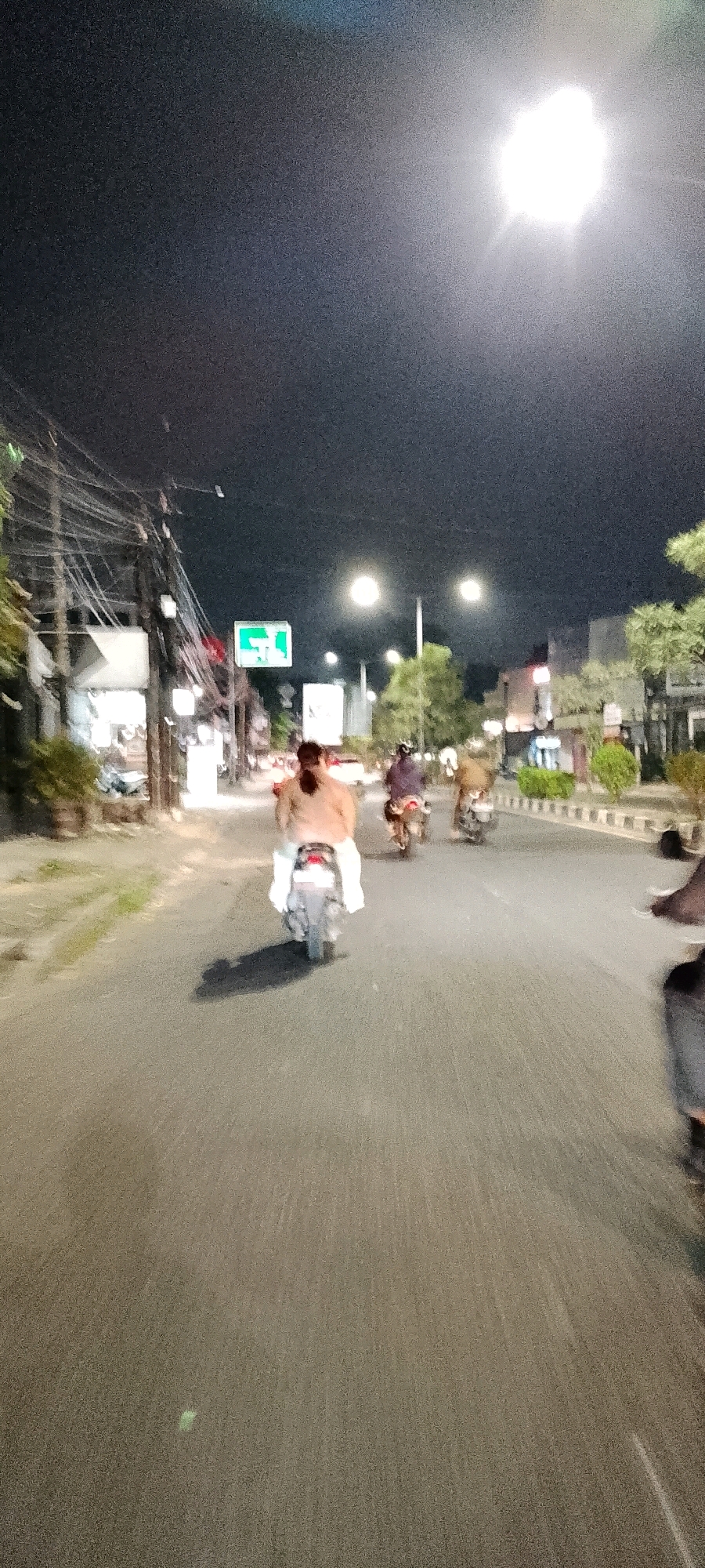 The image shows a nighttime scene on a street in Bekasi, Indonesia. The perspective is from the rear of a motorcycle, looking forward. A person wearing a light-colored garment is riding a light-colored motorcycle in the foreground. The person has long dark hair tied in a ponytail. To the right and slightly ahead of the main subject, two more motorcycles are visible, each carrying a rider. Further down the road, traffic lights and streetlights illuminate the scene. On the left side of the street, there are buildings with shops and a prominent green sign. Overhead, a dense network of electrical wires is visible. The road surface appears to be asphalt, and there are white road markings. The overall image is somewhat blurry due to motion and low light conditions.