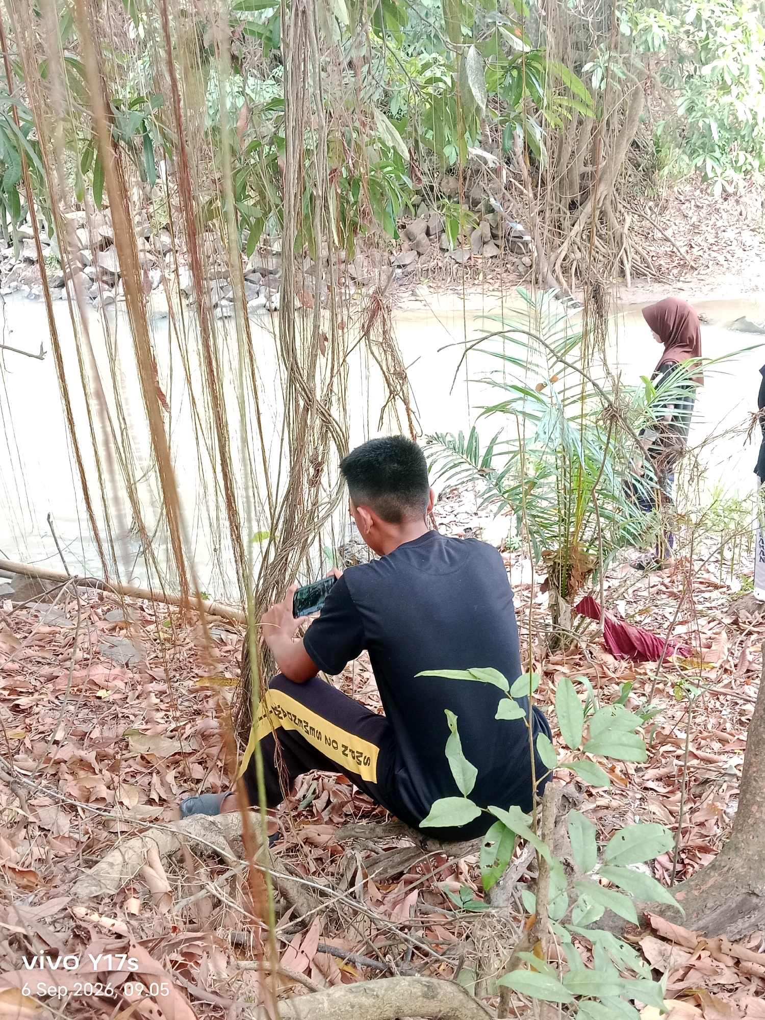The image depicts a riverside scene in Rama Kelandungan, Indonesia. In the foreground, a young man wearing a black t-shirt and black and yellow track pants is seated on the ground, facing away from the camera and towards the river. He is holding a mobile phone in his hands, seemingly engaged with it. Behind him and slightly to the right, another person, a woman wearing a brown hijab, stands near the riverbank. The environment is characterized by dense vegetation, with numerous hanging vines obscuring the view of the river and the background. The ground is covered in dry leaves and debris, with exposed tree roots and rocks visible. The river water appears light brown and shallow. The overall atmosphere suggests a natural, outdoor setting.