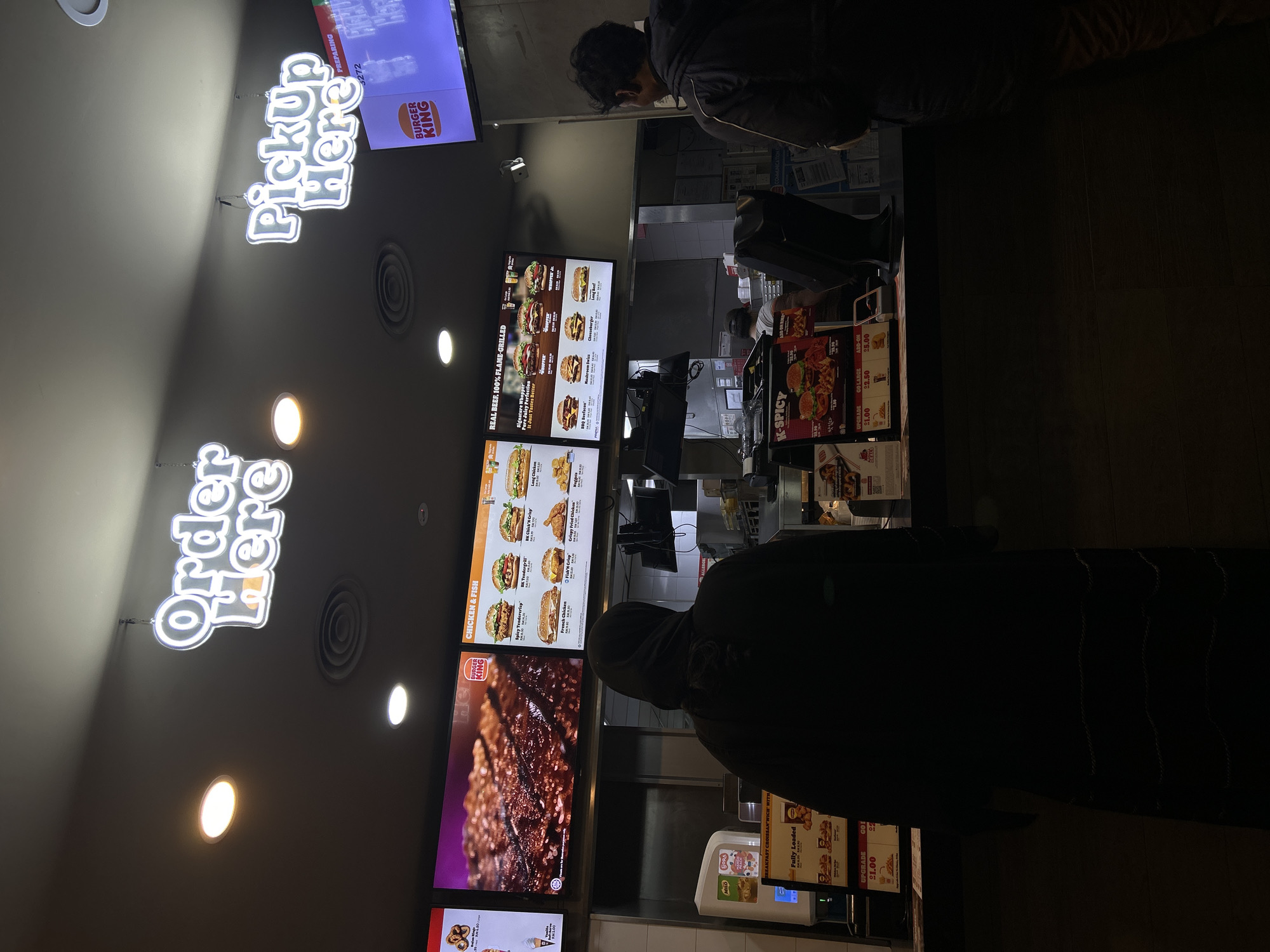 The image shows the interior of a Burger King restaurant. A customer is standing at the counter, seemingly placing an order. Digital menu boards display food items and prices. Signage for "Order Here" and "Pick Up Here" is visible in neon lights. There are menu boards that have words like "REAL BEEF. 100% FLAME-GRILLED" and "CHICKEN & FISH". The image was taken in Malaysia, Subang Jaya City Council.
