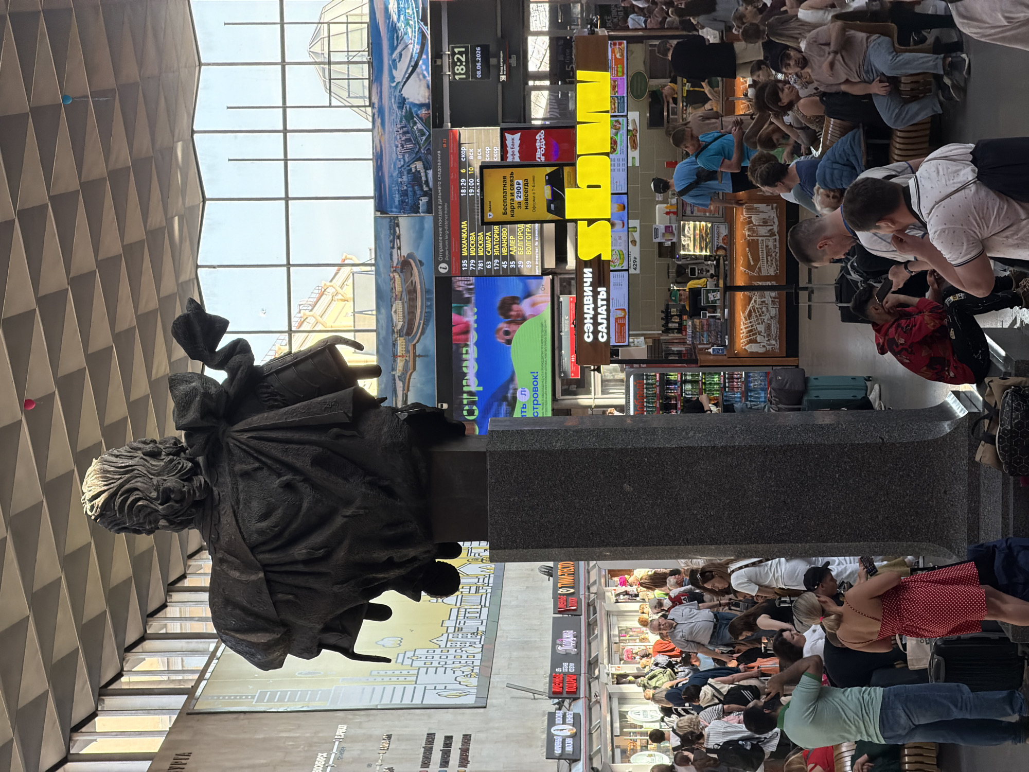The image captures a bustling interior scene, likely a train station or large public building, in Saint Petersburg, Russia. Dominating the foreground is a large, dark bronze bust of a historical figure, positioned on a tall, dark granite pedestal. The bust depicts a person with elaborate hair and flowing robes, holding what appears to be a piece of cloth or fabric in their left hand.
In the background, a crowd of people are seen standing and sitting, engaged in various activities. Some are looking at their phones, others are talking, and many are carrying luggage, suggesting travel. The environment features a high, patterned ceiling with triangular panels and large windows that allow natural light to filter in, revealing a clear sky outside.
Several digital display boards are visible, showing train departure information and advertisements. One prominent board displays "Отправление поездов дальнего следования" (Departure of long-distance trains) with a list of destinations and times. Another large screen shows colorful images and text. There are also various kiosks and shops, including one with prominent yellow signage that reads "subw," likely a food establishment. The overall atmosphere is one of transit and public gathering.