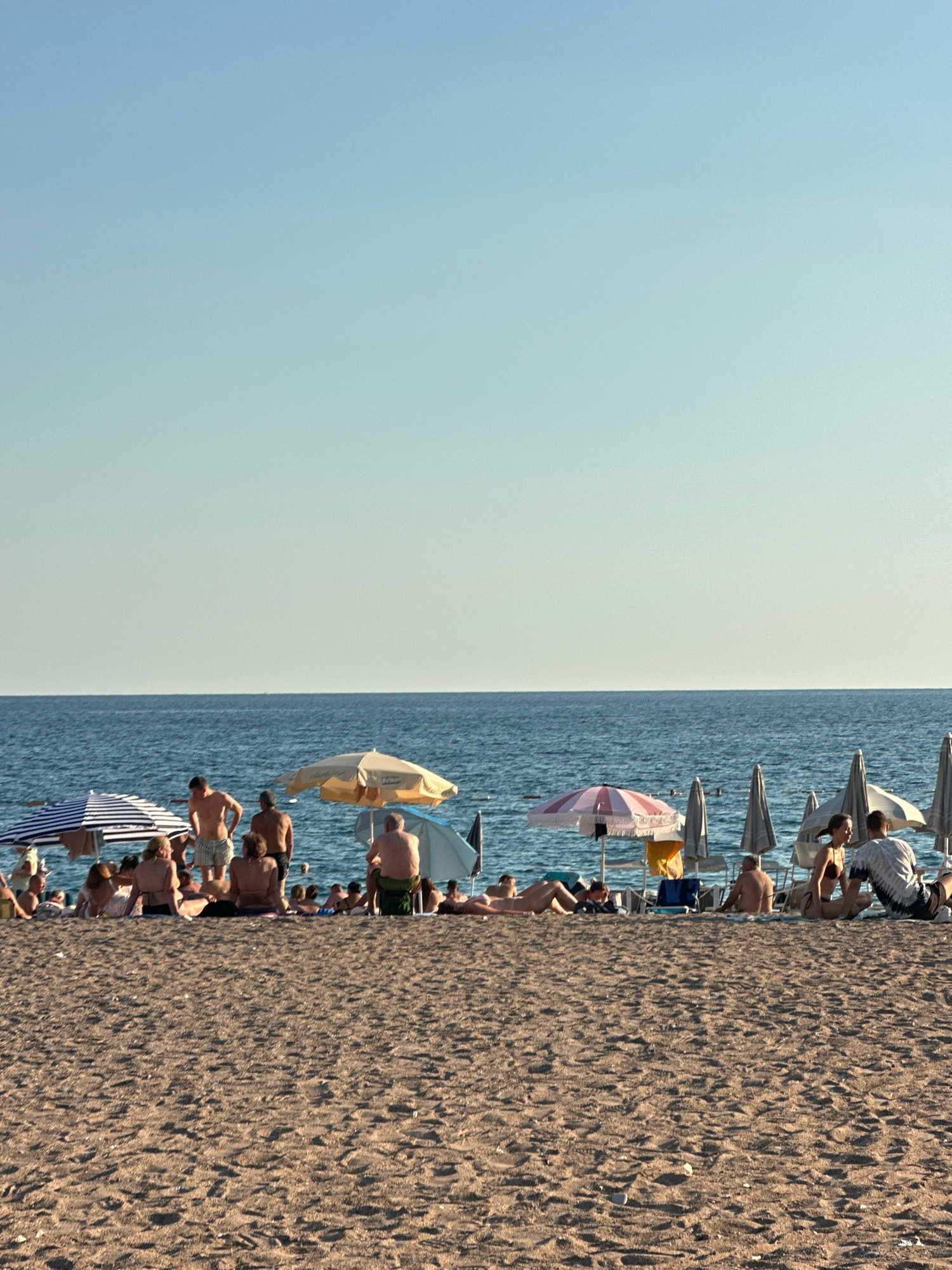 The image depicts a crowded beach scene on a sunny day, likely in Boreti, Montenegro, given the context. The foreground is dominated by granular sand, showing footprints and scattered pebbles. In the mid-ground, a collection of beachgoers are situated on the sand, many reclining or sitting. Several beach umbrellas, some striped and others solid-colored, are erected to provide shade. The water, a deep blue, stretches across the background, meeting a clear, pale blue sky at the horizon. People are seen in various states of relaxation, some standing, others seated or lying down, with many wearing swimwear. The overall atmosphere is that of a typical summer day at a popular coastal resort.