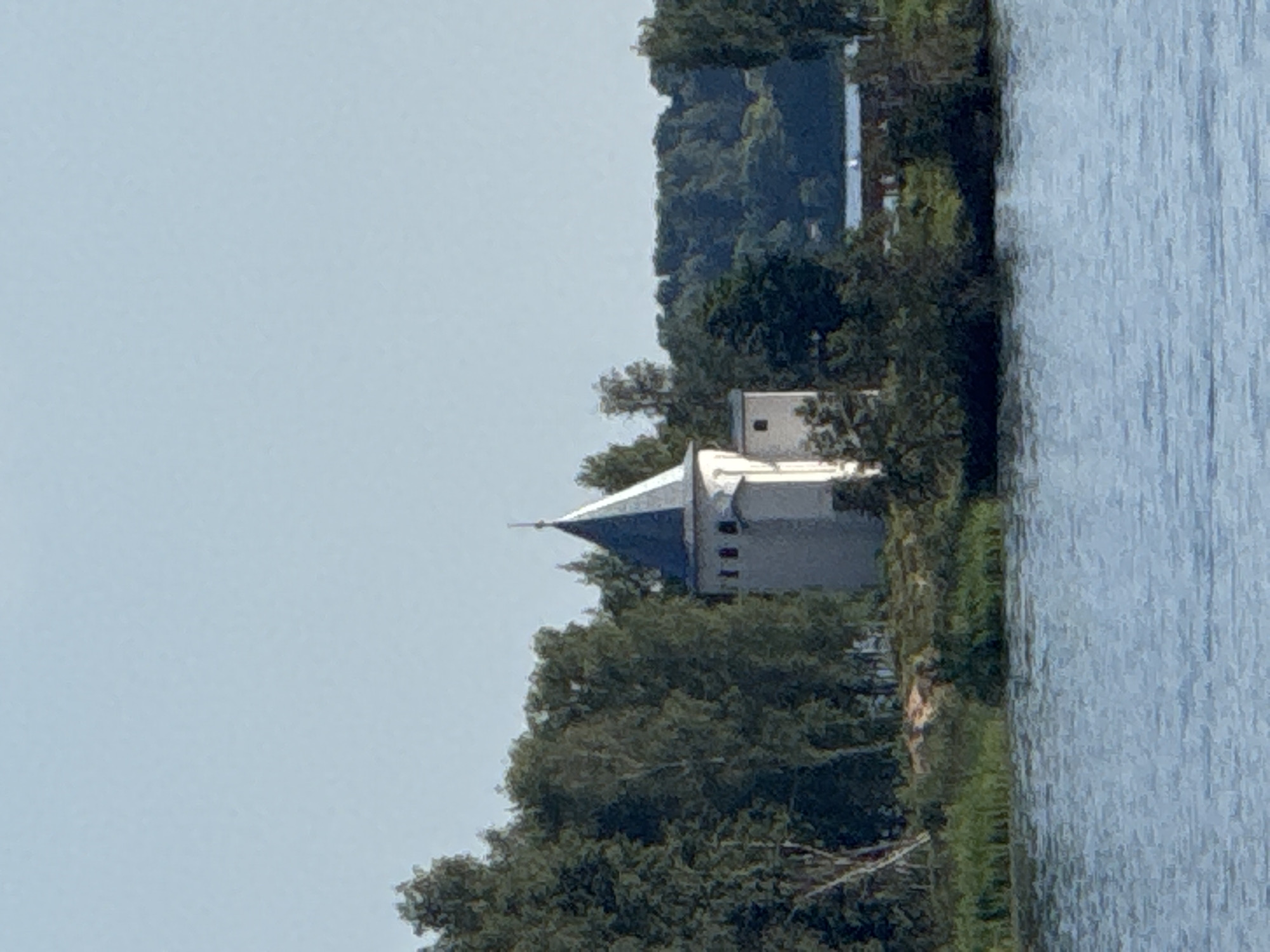 The image captures a serene view of a white tower with a dark gray, conical roof, situated on a treelined shore. The tower appears to be part of a historical complex, possibly a church or monastery, with small windows visible on its upper section. Lush green trees surround the tower, creating a natural backdrop. In the foreground, a calm body of blue water reflects the sky and the surrounding greenery. The overall scene suggests a peaceful, possibly rural, setting. This structure is likely the bell tower of the flooded St. Nicholas Monastery in Kalyazin, Russia, a prominent landmark that partially remains above the Ivanovo Reservoir.