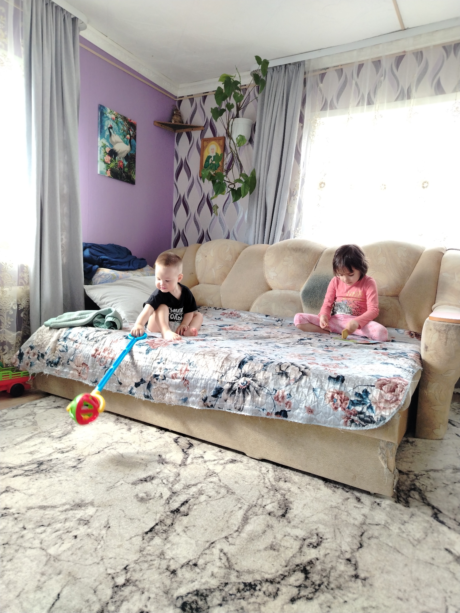 A photograph taken in what appears to be a residence in Гожан, Russia, shows a young boy in a black shirt playing with a toy on a large sofa while a girl in pink sits nearby. The sofa is covered with a floral-patterned quilt. Behind them, the wall is purple with patterned wallpaper and a framed image. To the left, a curtain partially obscures a window, while to the right, another window with sheer curtains lets in bright daylight. A potted plant is positioned near the window on the right. The floor is covered with a marble-patterned rug.