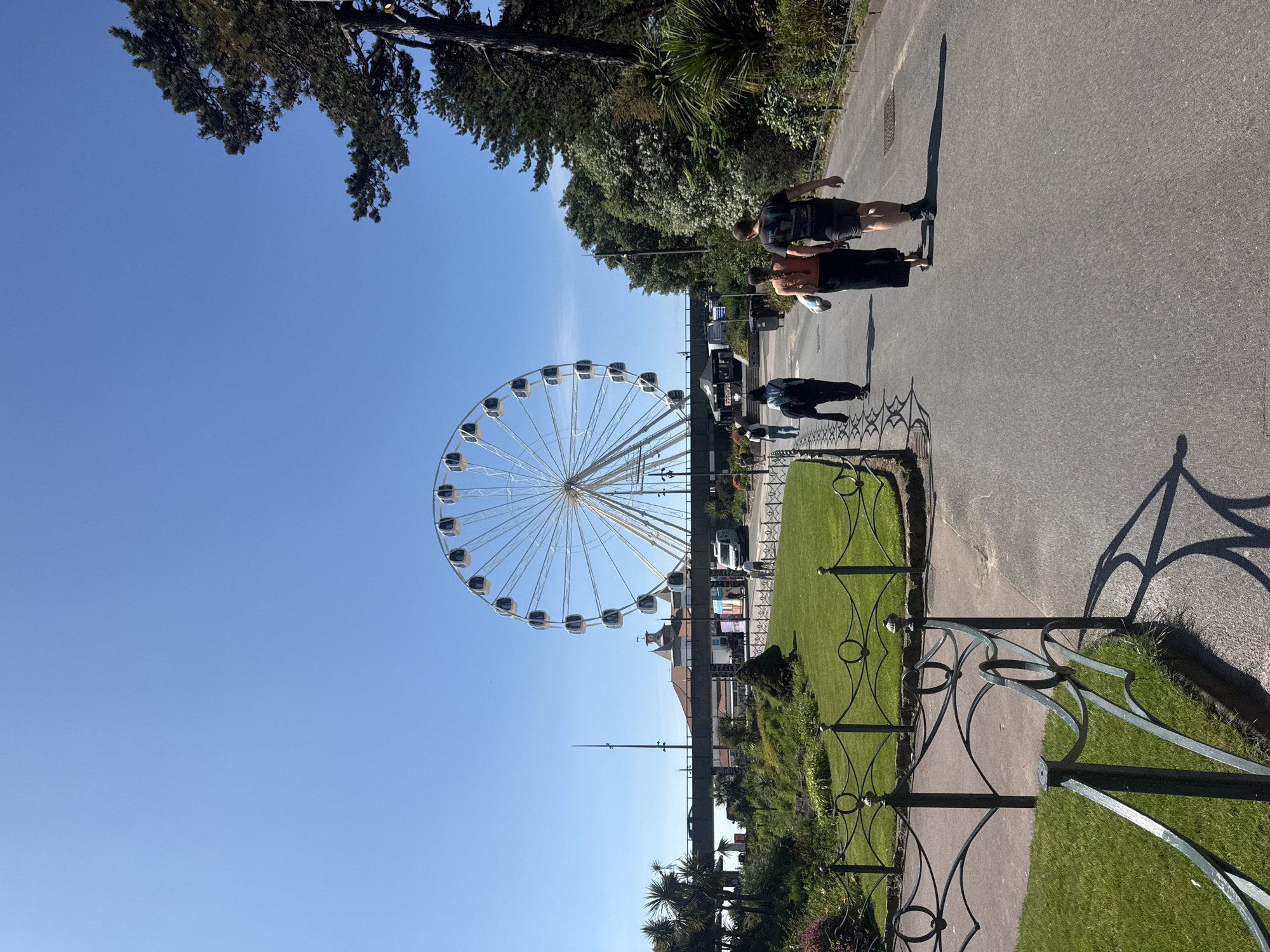This is a vertical, eye-level, full shot of a Ferris wheel and a park in Bournemouth, United Kingdom, on a clear day with a bright blue sky. The Ferris wheel is in the background, to the left of center, and is a large white structure with many gondolas. In front of the Ferris wheel, there is a walkway with several people walking. To the left of the walkway, there is a grassy park area with a decorative fence. To the right of the walkway, there are trees and foliage. In the far background, there is a bridge and some buildings. The overall scene is one of a sunny day at an amusement park or tourist attraction.
