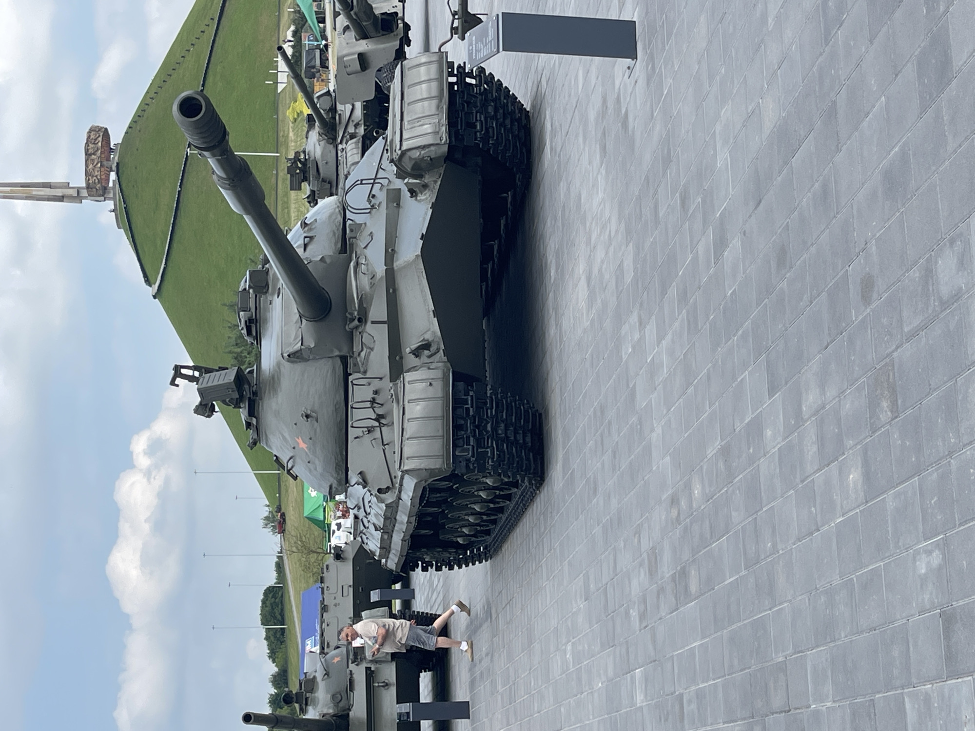 The image shows a large, grey tank displayed outdoors on a paved area. The tank is the primary subject, with its turret and tracks clearly visible. In the background, a grassy hill slopes upwards, topped by a monument. The sky is cloudy, suggesting it might be daytime with overcast weather. A person in shorts and a t-shirt is standing near the tank, appearing to observe it. To the left of the tank, the environment includes trees, poles with lights, and tents, indicating an outdoor exhibition or museum setting. The scene appears to be a memorial or a military museum due to the presence of the tank and monument. No specific text is clearly legible in the image.
