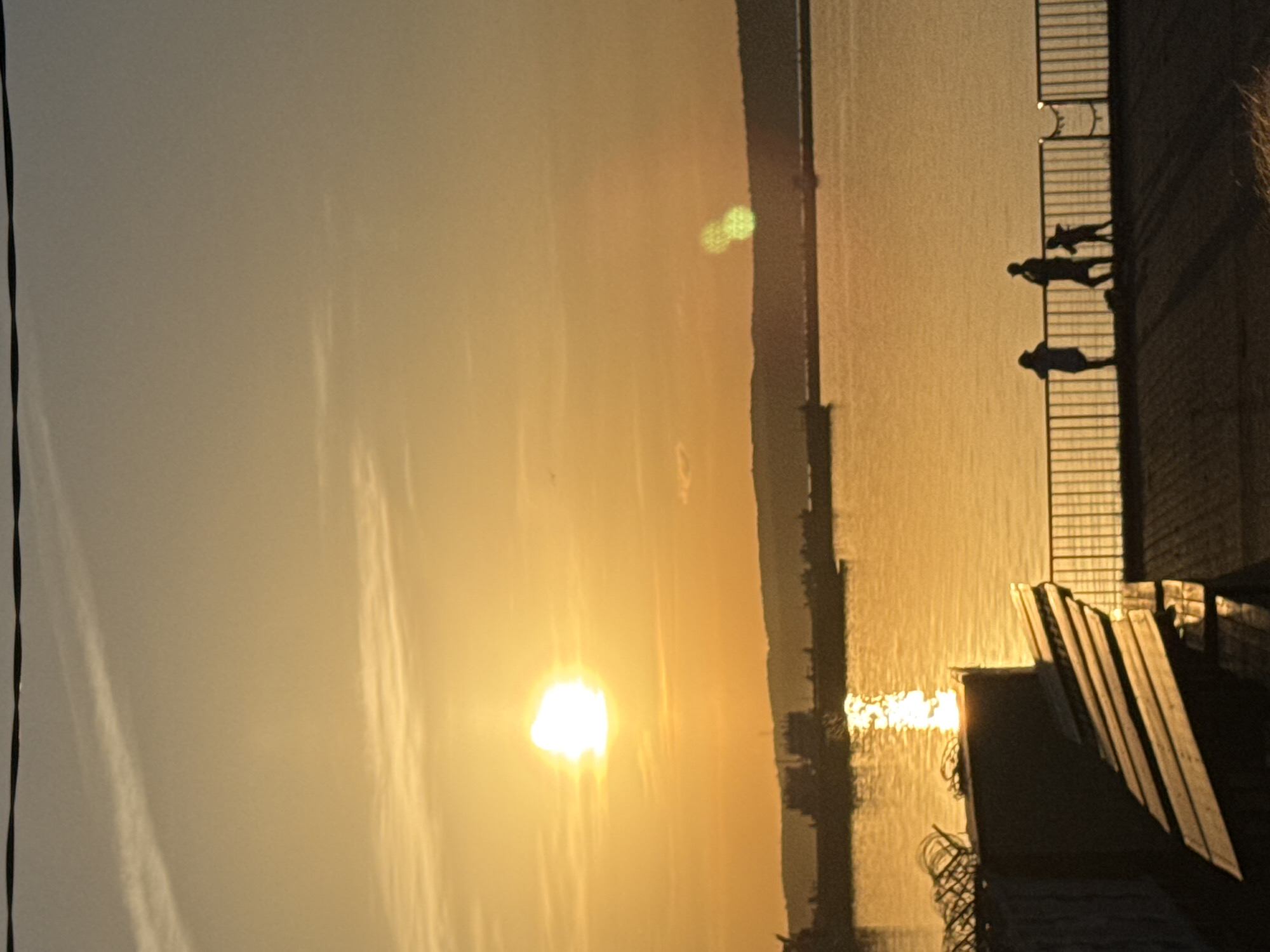 Captured near Chaikovsky, Russia, the image shows a golden sunset over a body of water. Silhouetted figures stand along a railing, seemingly observing the sunset. The landscape across the water is dimly visible, adding depth to the scene. Structures and building elements are also silhouetted in the foreground.