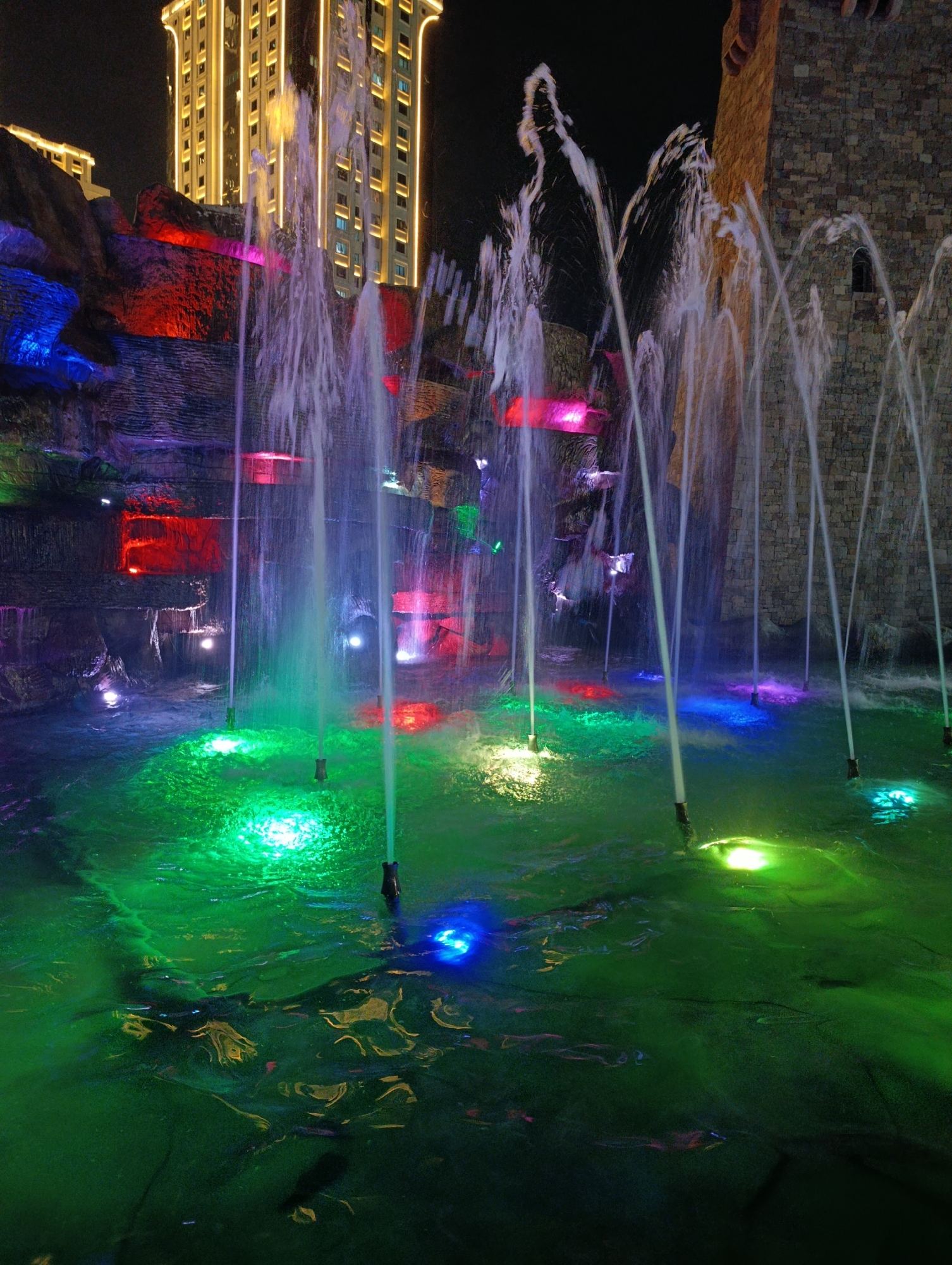 The image shows an outdoor night scene featuring a water fountain illuminated by various colored lights. The fountain sprays water upwards in several jets, creating a dynamic visual effect. The surrounding environment includes a textured rock formation with embedded colored lights, a stone tower to the right, and a tall, modern building in the background with numerous illuminated windows. The water in the pool below the fountain is reflecting the lights, creating a vibrant green hue with patches of blue and pink. The scene appears to be a decorative public space, possibly a park or plaza, designed for evening entertainment. Based on the architectural style of the background building and the known landmark, this scene is located in Grozny, Russia, likely at the "Grozny-City" complex. There are no people visible in the image.
