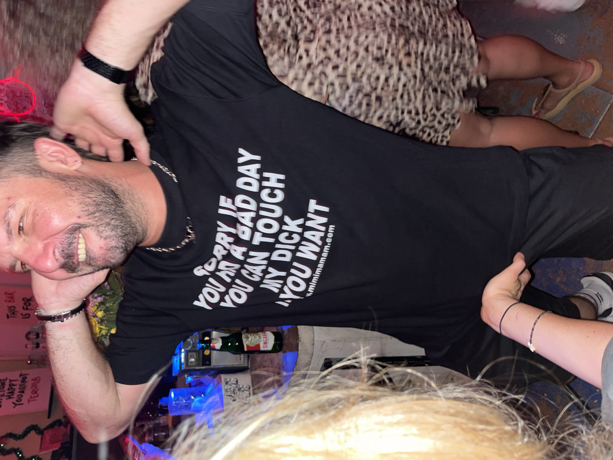 A bar in North Kuta, Indonesia. A man with grey-flecked dark hair and beard is smiling, wearing a black T-shirt with the words "SORRY IF YOU HAD A BAD DAY YOU CAN TOUCH MY DICK YOU WANT" and "mimimamam.com" on it. A woman wearing a leopard print dress and beige sandals is standing behind him. Another person is standing beside him with their arm touching his hip. There is a sign reading "THIS BAR IS FOR HAPPY YOU ARE HOT TERULA" next to the man. A bottle of "SINGARA" beer is also visible. The bar appears to be decorated with festive lights and decorations.