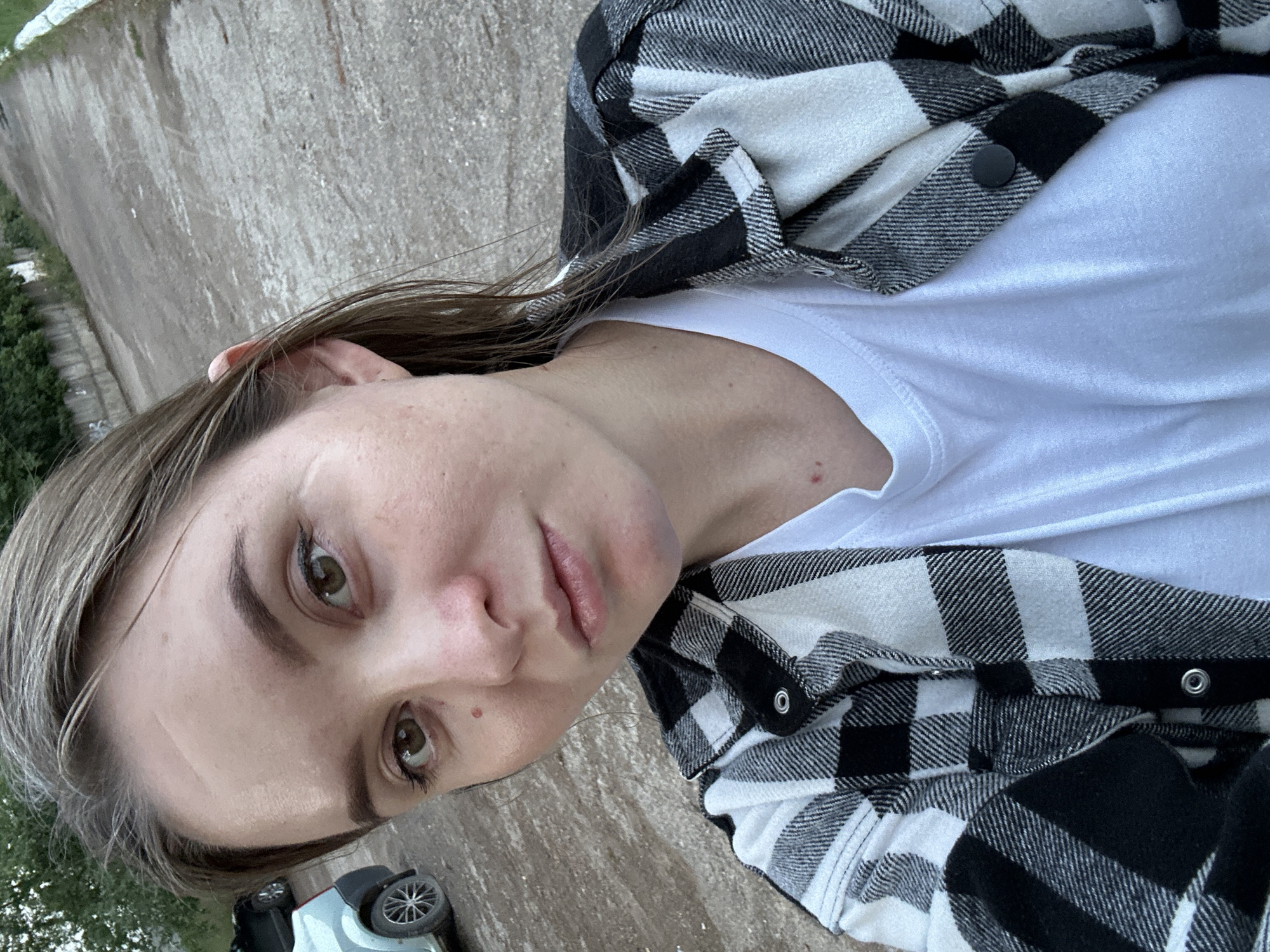 A young woman with brown hair is taking a selfie outdoors. She is wearing a white t-shirt and a black and white plaid shirt. Her eyes are open, and she is looking directly at the camera. The background appears to be a street or road with some trees and a concrete wall. There are two vehicles visible in the background, one a light-colored car, the other a dark-colored vehicle partially obscured. The ground is paved and appears to be damp or dirty. The lighting suggests it is daytime, possibly overcast. The location is identified as Noviy, Russia.