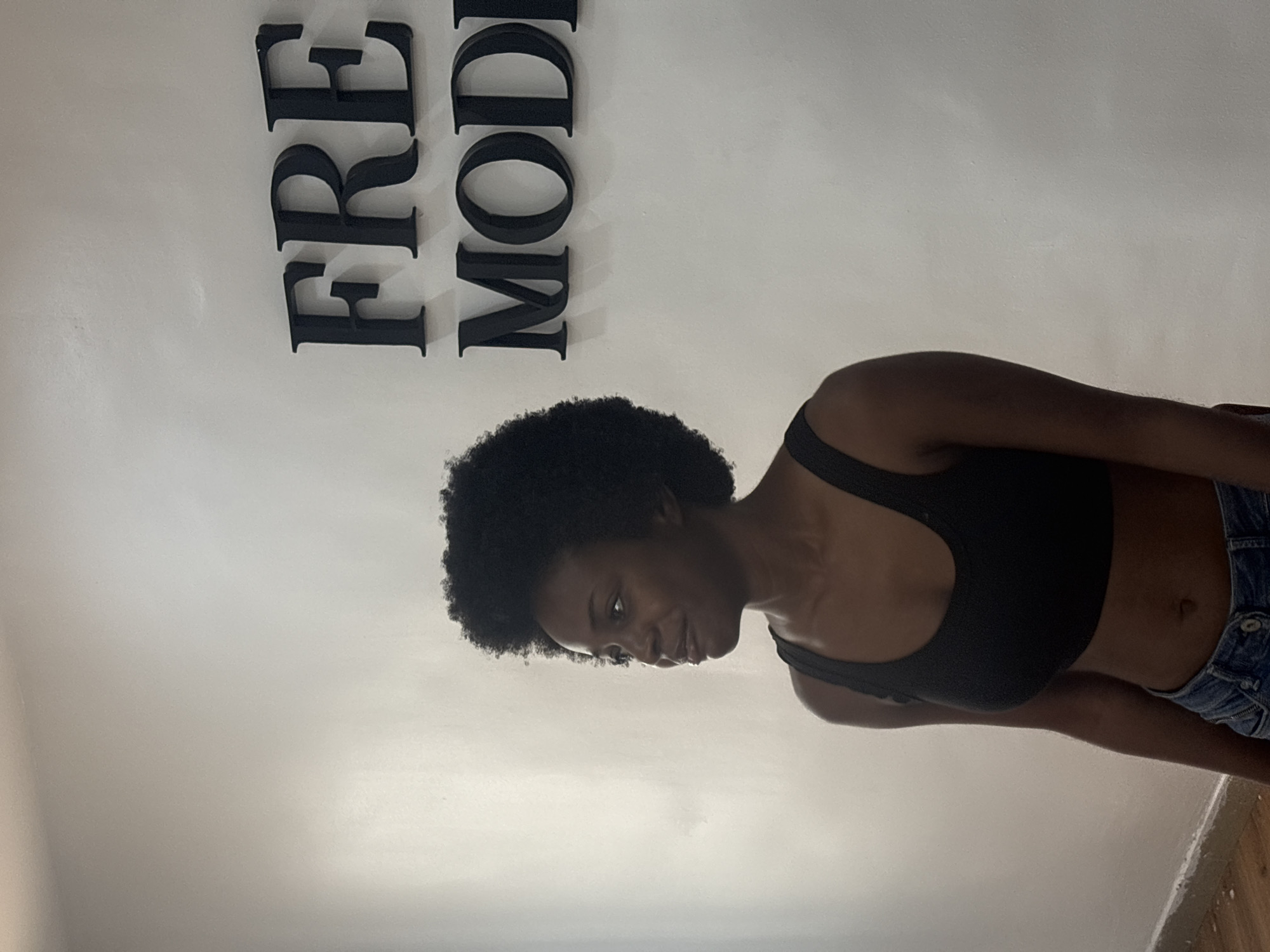 A young, dark-skinned woman with an afro is standing in front of a white wall with black lettering that reads "FREE MOD". She is wearing a black sports bra and blue denim shorts. The location is likely in Lagos, Nigeria, based on the lettering. The image seems to be taken in an indoor environment.