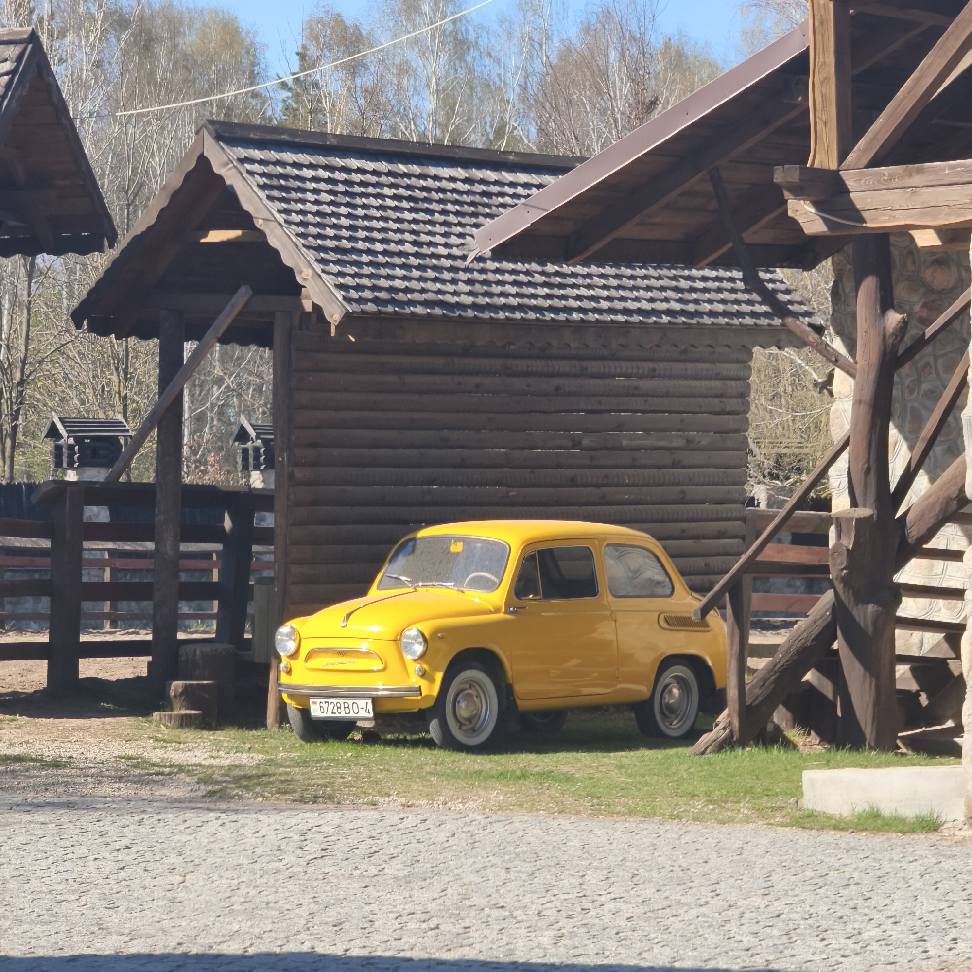 The image depicts a yellow vintage car, parked in front of wooden buildings with distinctive roofs in Капцёўскі сельскі Савет, Belarus. The car's license plate is visible and reads "6728 BO-4". The building behind the car has a dark shingled roof. Another wooden structure with a different roof design is visible to the left of the car. The foreground is a stone-paved area. Bare trees can be seen in the background.