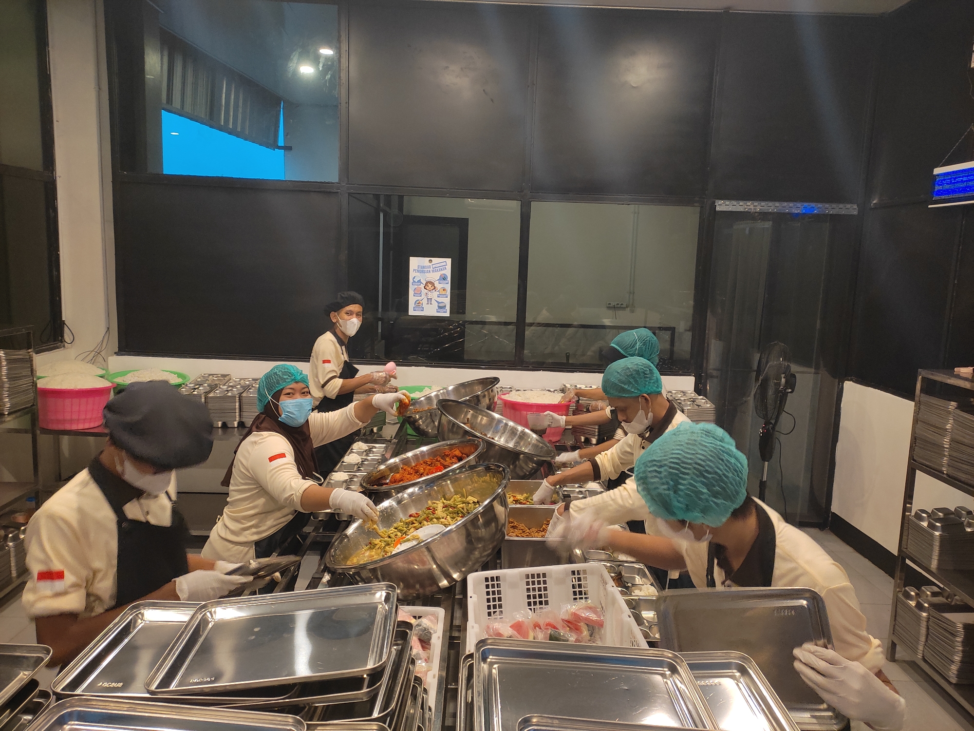 In a kitchen environment in Ponorogo, Indonesia, several individuals wearing hairnets and masks are actively preparing food. The scene appears to be a catering or food service operation. Several large metal bowls filled with various dishes are visible, including what looks like rice, a stir-fry of vegetables, and a reddish-brown saucy dish. People are using serving utensils to portion food into smaller metal containers and trays. Stacks of clean metal trays and containers are present on shelves and work surfaces, indicating organized food preparation and service. The overall atmosphere suggests a busy food preparation area.