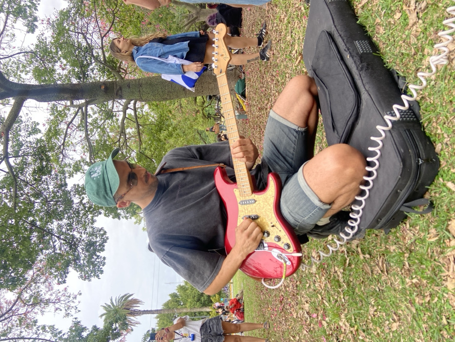 This image depicts a male musician kneeling and actively playing a red electric guitar in the Bosques de Palermo, Buenos Aires, Argentina. The scene occurs on a grassy area with scattered dry leaves, under an overcast sky, surrounded by mature trees including a distinct *Palo Borracho* (floss silk tree) with a textured trunk and a palm tree visible in the background.
The primary subject, a male in his 20s or 30s, wears a green baseball cap, dark sunglasses, a dark grey short-sleeved t-shirt, and blue denim shorts. He is supported by his knees resting on a black soft guitar case placed on the grass. He plays a red electric guitar, possibly a Fender Stratocaster, characterized by its light-colored, possibly pearloid, pickguard and three single-coil pickups. A white coiled instrument cable is connected to the guitar's output jack and extends towards the ground. A brown leather strap supports the guitar.
In the mid-ground, a female stands to the right, wearing a denim jacket over a black dress and carrying a white tote bag with blue markings. To the left, a partial view of another female shows her holding a smartphone, seemingly recording the event. Additional indistinct figures are scattered throughout the park in the background, indicating a public gathering or leisurely activity.