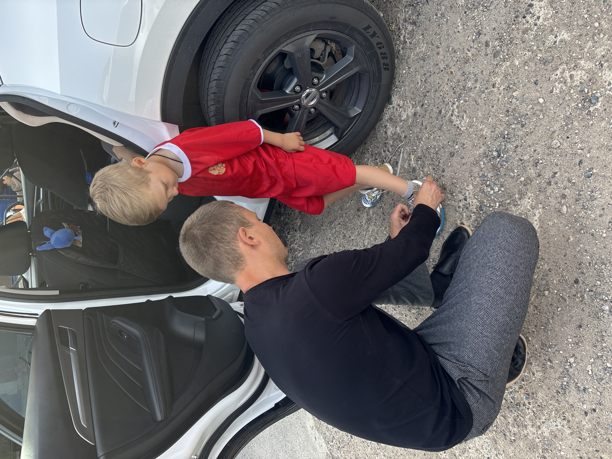 This media file shows a man and a young boy kneeling on a gravel surface next to a white car. The man, wearing a black long-sleeved shirt and checkered gray pants, is assisting the boy with his shoelaces. The boy, dressed in a red soccer jersey with white trim and a crest, is looking down at his feet. A portion of the car's interior, including a black leather door panel and seats, is visible. The car's tire, with the text "LY688" on its sidewall, is also prominent. The scene suggests a casual, outdoor setting, possibly a driveway or parking area, given the gravel ground and the car. The lighting indicates it is daytime. The interaction between the man and the boy appears caring and attentive. There is no indication of specific weather conditions.