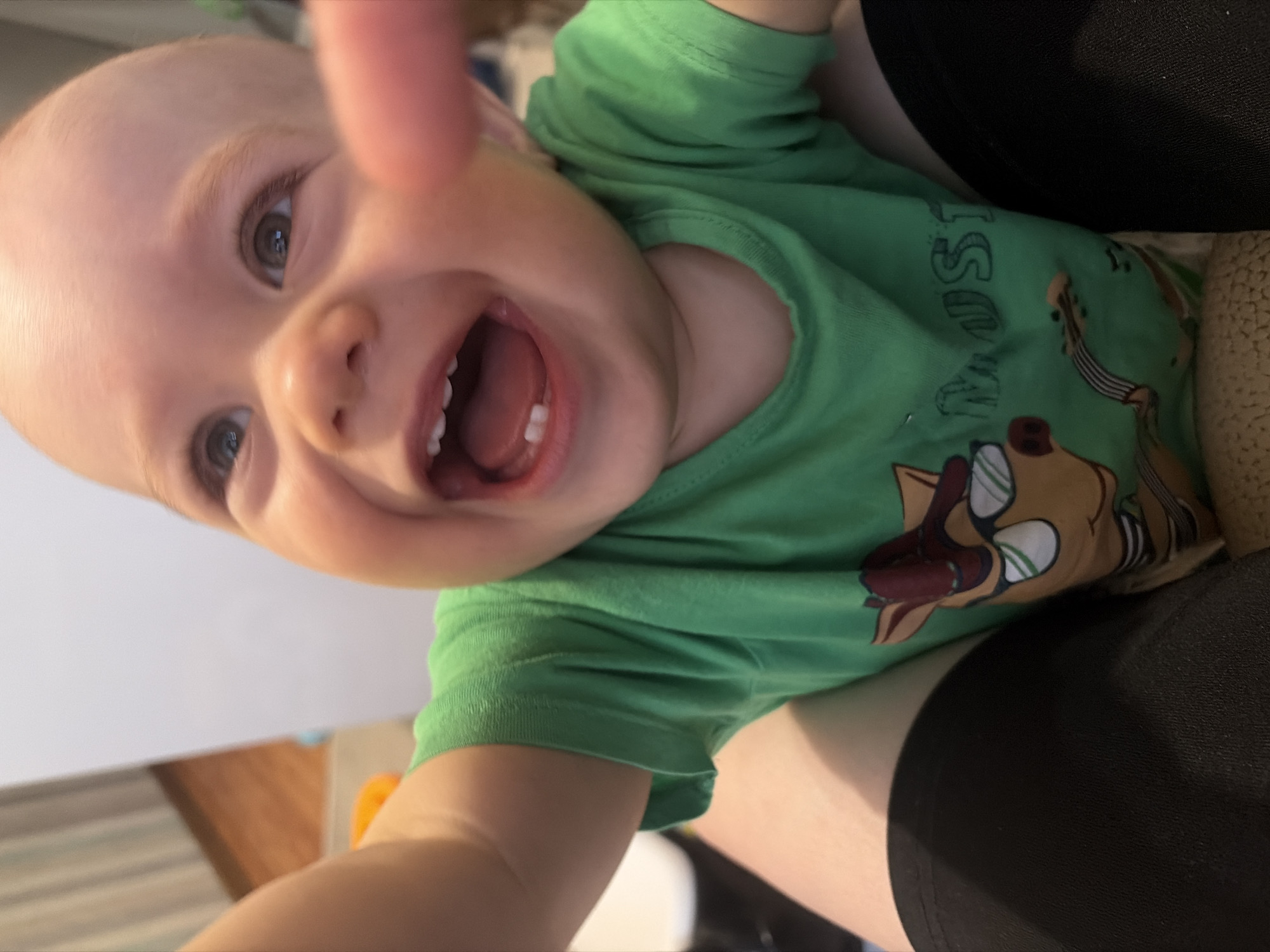 A fair-skinned baby wearing a green t-shirt with a cartoon graphic is being held. The baby has a wide, toothy smile, light hair, and gray-blue eyes. Part of an adult's arm and black-clothed lap are visible holding the baby. A finger is partially in the frame. The background includes a white surface and a glimpse of a wood-toned floor. The location of the image is unknown.