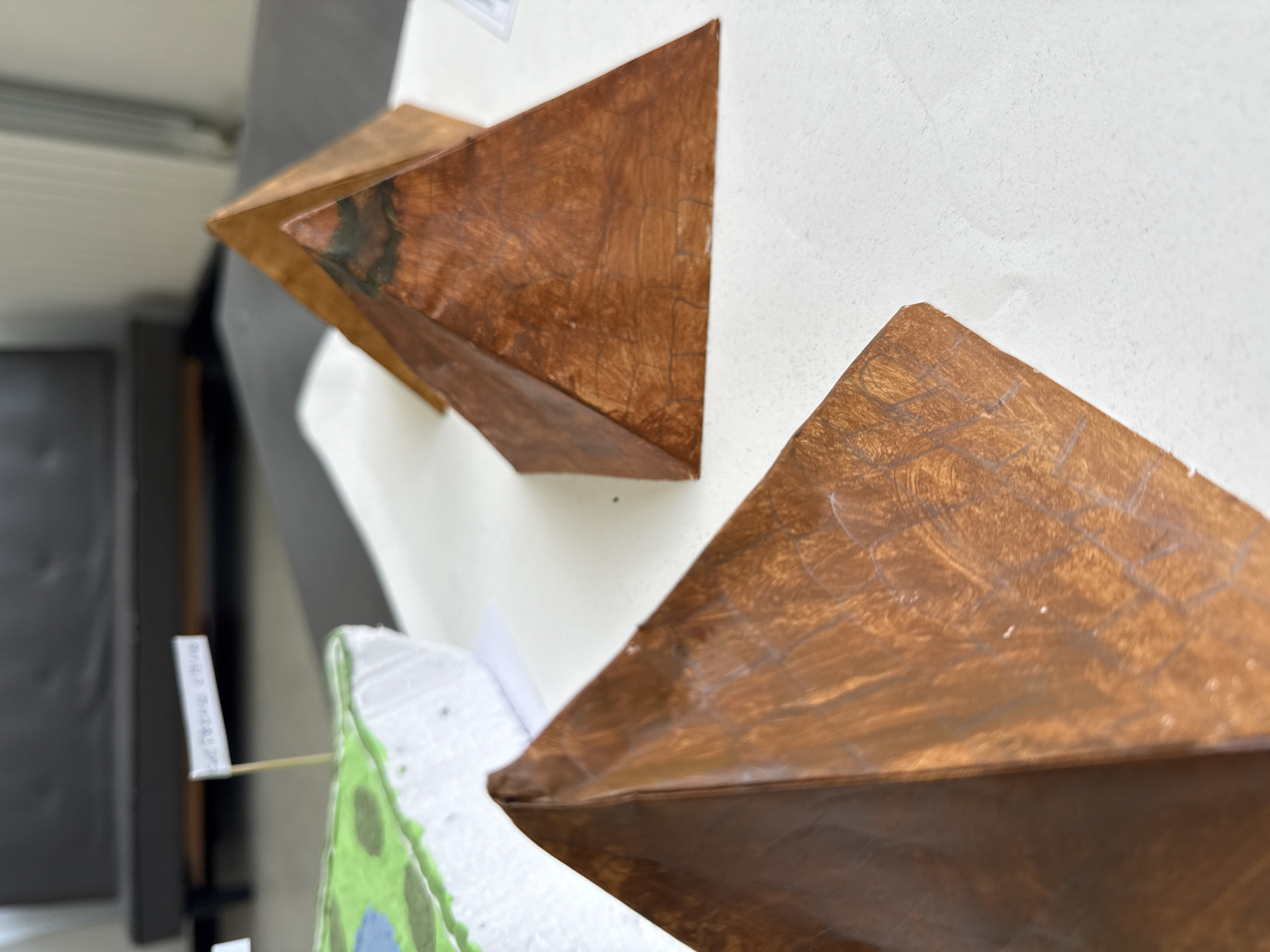 The image displays an indoor scene featuring several geometric models arranged on a white surface, possibly a table or display board. Three prominent pyramid-like or prism-like shapes, likely constructed from cardboard or a similar material, are painted a reddish-brown color, giving them a textured, wood-like or metallic appearance. Some surfaces exhibit faint lines, possibly construction guidelines, and one of the shapes has a notable dark greenish-blue area on one of its faces.
In the bottom left corner, a portion of another model is visible. This model appears to be a landscape or biological structure, made from a white, textured material (possibly styrofoam) with painted green and blue sections. A small white flag, attached to a toothpick, is inserted into this model. The text on the flag is blurred and illegible.
The background includes a dark, blurred area to the left, which might be a piece of furniture or another display element, and a white wall or screen behind the models. The arrangement suggests an educational exhibition or a school project display. No specific activity is taking place; the models are static exhibits. The lighting appears to be ambient, typical of an indoor setting.