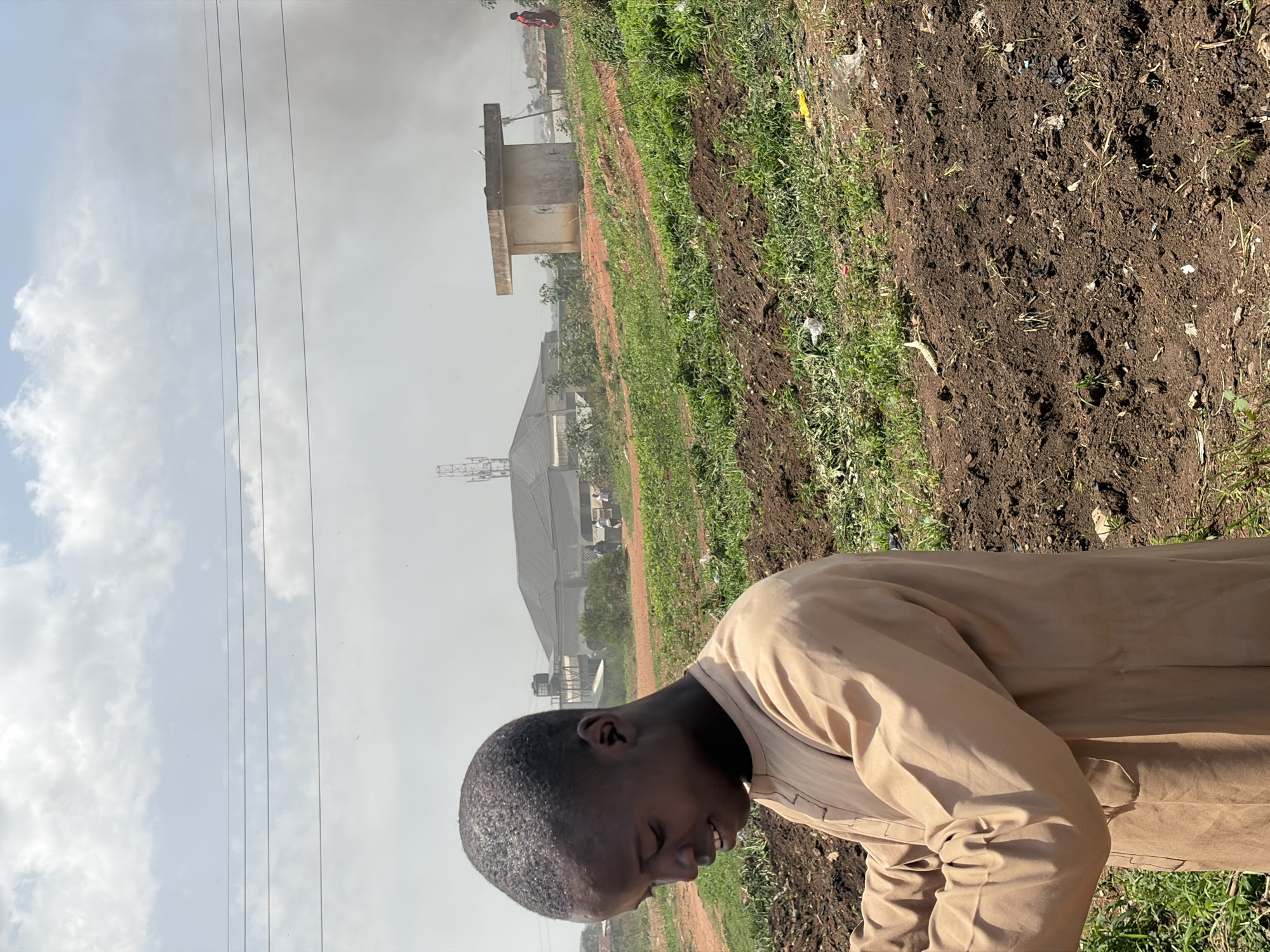 An eye-level, medium shot of a young boy, likely of African descent, wearing a light-brown robe. The boy is in profile, looking down with his head angled slightly to the left. The location is the city of Barakin Lerawi, Nigeria. The background includes a field of grass and dirt, utility poles and power lines against a cloudy sky, and a distant building with a cell tower. A concrete structure is also visible in the midground. The boy is in the foreground and appears to be looking at the ground or something near his feet.