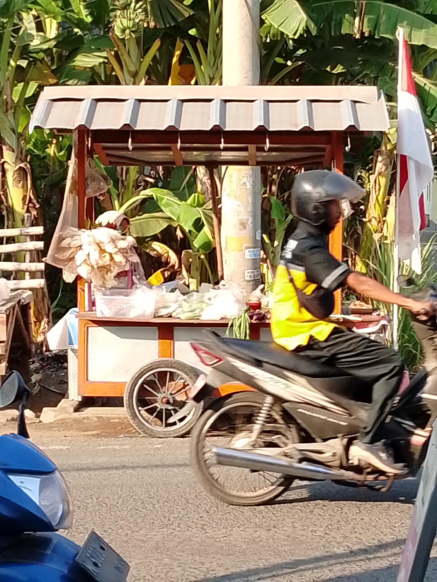 In Pasuruan, Indonesia, a motorcycle rider wearing a black helmet and a yellow and black uniform rides on a paved road. They are passing a roadside stall with a brown and orange frame, covered by a light-colored corrugated roof. The stall displays various items, including what appears to be produce wrapped in plastic. Behind the stall, a variety of lush green banana trees are visible. To the right of the stall, a red and white Indonesian flag is flying. In the foreground, the front of a blue motorcycle is partially visible, indicating another vehicle is present. The scene is illuminated by bright sunlight, casting shadows on the road and stall.