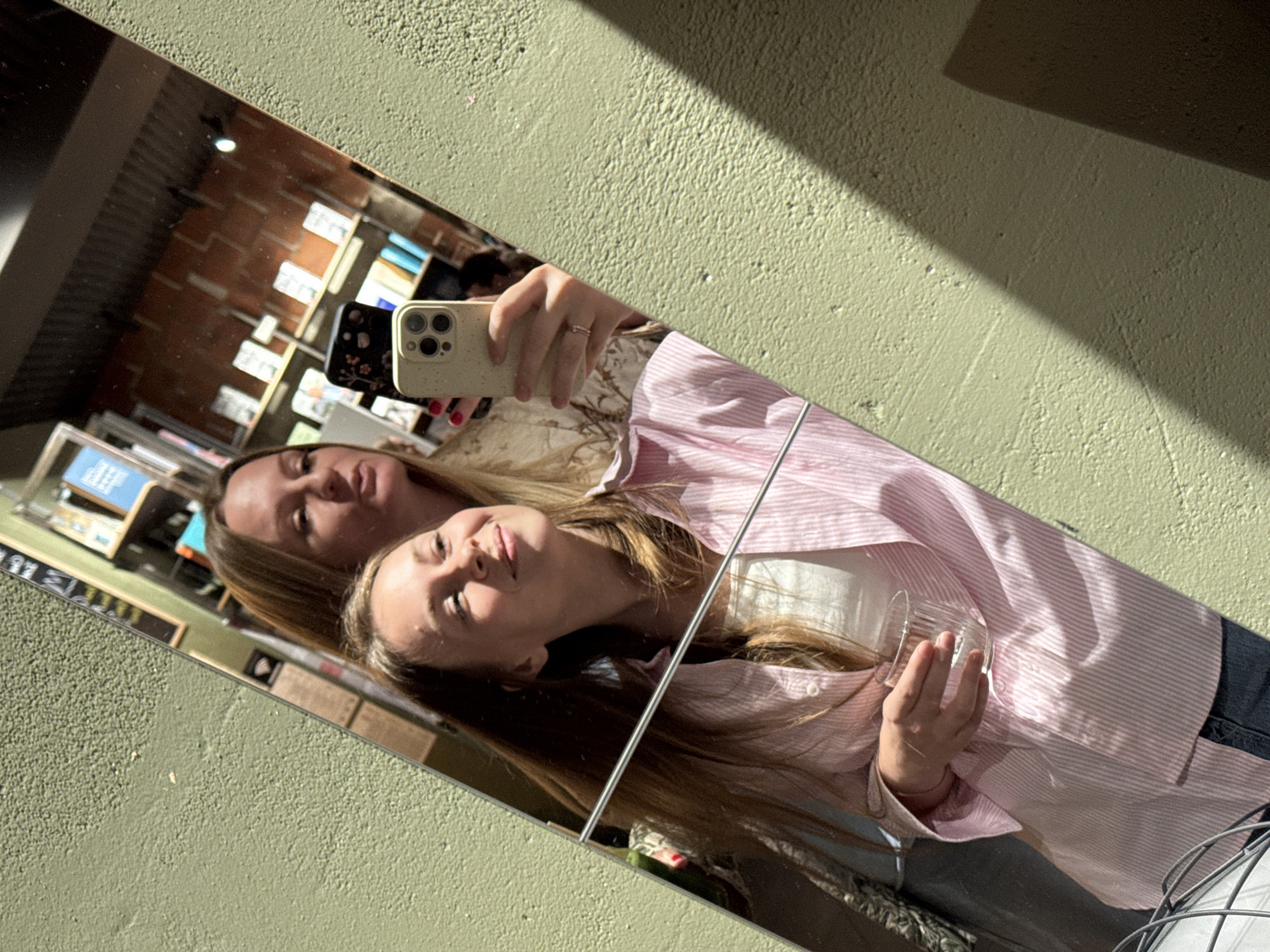 Two women are reflected in a mirror, with the mirror positioned against a textured, light green wall. The reflection reveals an interior space, possibly a store, with shelves stocked with items. One woman holds up a tan-colored iPhone to take the photo, while the other is positioned behind her. The woman in front is wearing a pink and white striped shirt, and she holds a small glass. The other woman is wearing a patterned shirt. The location is possibly a store in Budva, Montenegro, given the prompt.