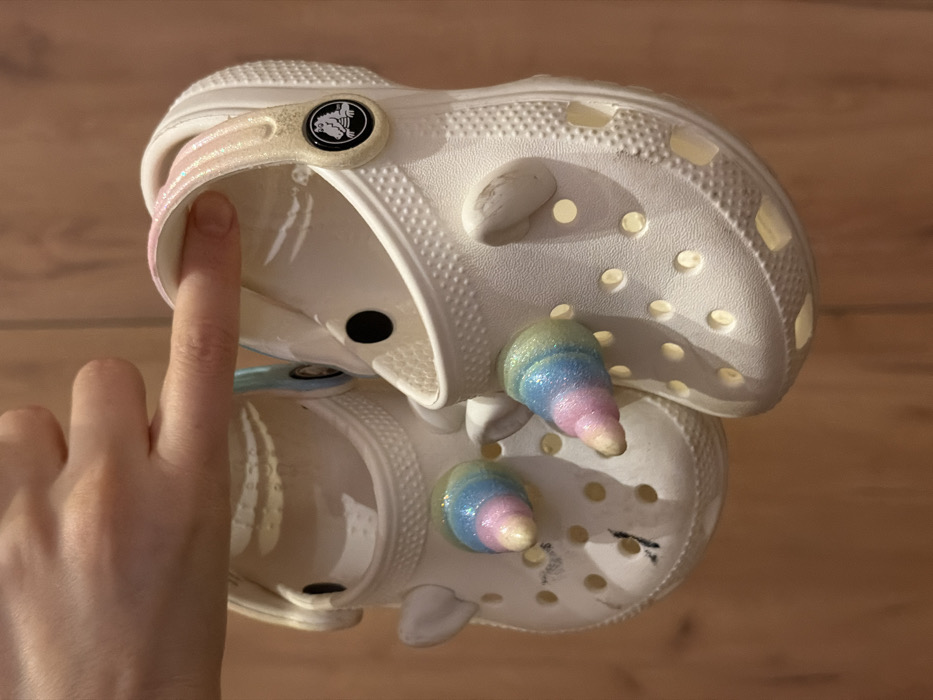 A human hand, characterized by a light skin tone, suspends two white Crocs brand clogs against a light brown wooden panel background in Rzhavki 2, Russia. An index or middle finger passes through the glittery light pink and gold strap of the right clog, while a thumb rests against the strap of the left clog. Both clogs are adorned with two iridescent, glitter-coated unicorn horn Jibbitz charms, featuring a blue, pink, and yellow gradient, along with small white ear-shaped charms. The right clog displays the black and white Crocs alligator logo on its strap button. The left clog exhibits dark scuffing or marks on its upper front surface. The background consists of vertically oriented wooden planks.