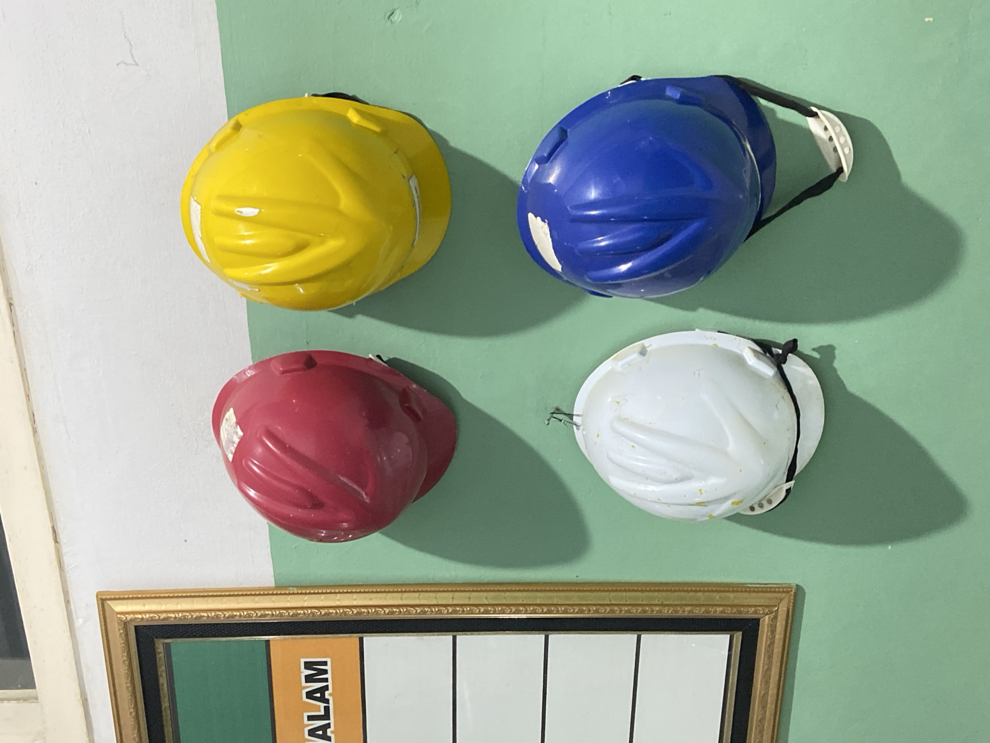 A collection of four hard hats is displayed on a wall, each suspended by a strap. The hard hats are in four different colors: yellow, blue, red, and white. They are arranged in a 2x2 grid. The yellow and blue hard hats are in the top row, with the yellow on the left and the blue on the right. The red and white hard hats are in the bottom row, with the red on the left and the white on the right. The wall is painted in two different colors, white on the left and a light green on the right. A framed picture hangs horizontally below the hard hats. The picture frame is gold with decorative detailing. Below the frame, a portion of a chart or poster is visible with vertical black lines and some text. The word "ALAM" is visible in large, bold, orange lettering on a darker background, part of the poster. The lighting suggests it could be daytime, with shadows cast by the hard hats indicating a light source from the left. The overall scene appears to be indoors, possibly a storage area or workshop, in Margoyoso, Indonesia. There are no people visible in the image.