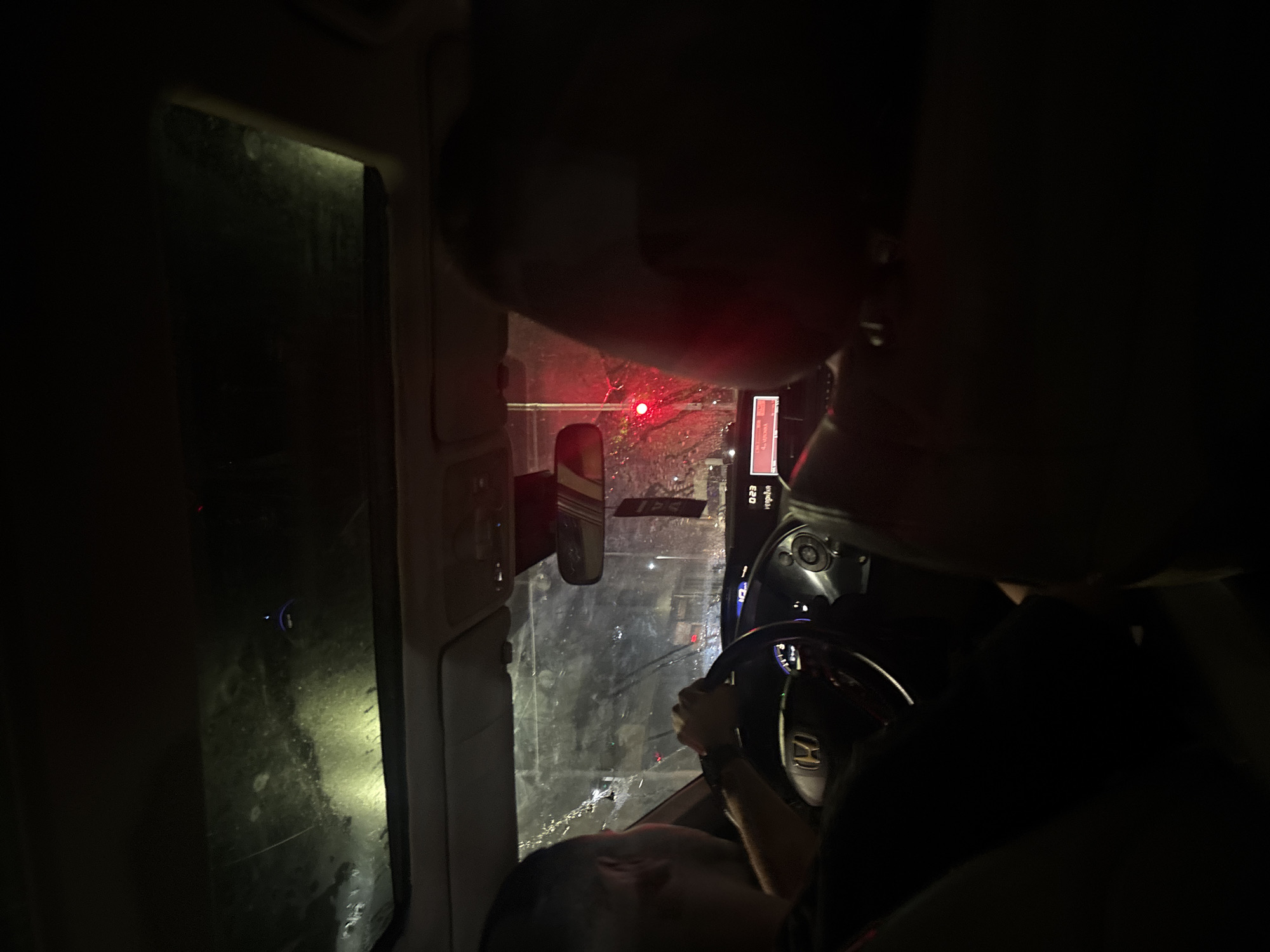 A nighttime, interior shot from inside a vehicle reveals a person driving, their hands gripping the steering wheel. Rain streaks the windshield, and the panoramic sunroof above shows a dark, wet sky. The car's dashboard displays "0:23," indicating the time. A red traffic light is visible outside, suggesting the vehicle is stopped at an intersection. The scene is dark, with the interior lights of the car offering minimal illumination. The setting appears to be a city street in Plovdiv, Bulgaria, characterized by wet roads and urban infrastructure reflected in the glass.