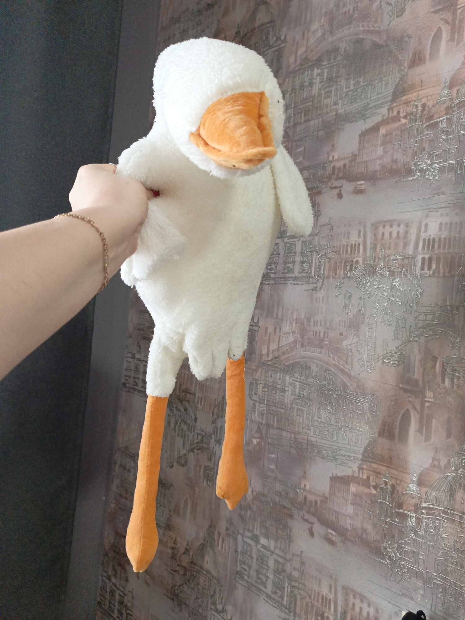 A person's hand, adorned with a gold bracelet, holds a white plush toy goose with orange legs and beak. The goose is positioned against a wallpaper featuring a faded, sketched cityscape, reminiscent of Venice. The scene is indoors, with a dark gray wall partially visible to the left. The overall impression is a casual snapshot of a personal item. There is no visual confirmation that the location is in the city of Марковский, Russia.