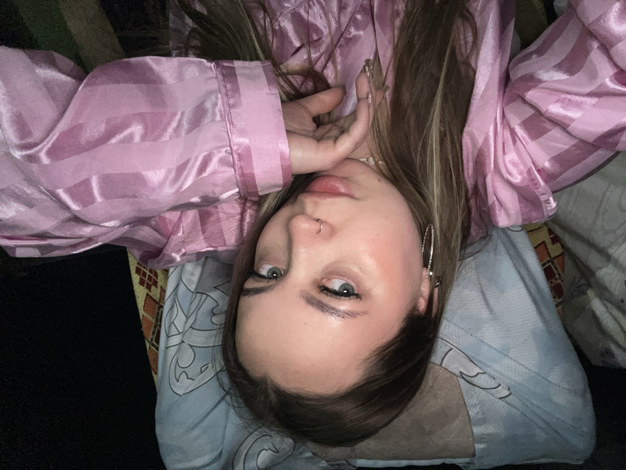 A young woman lies on a pillow, wearing a pink striped satin pajama top. She has long brown hair with blonde highlights, light skin, and is wearing hoop earrings. Her nails are long and decorated. The background includes parts of other pillows or bedding. There is no evidence to suggest the location is in Novopokrovskaya, Russia, or any city at all.